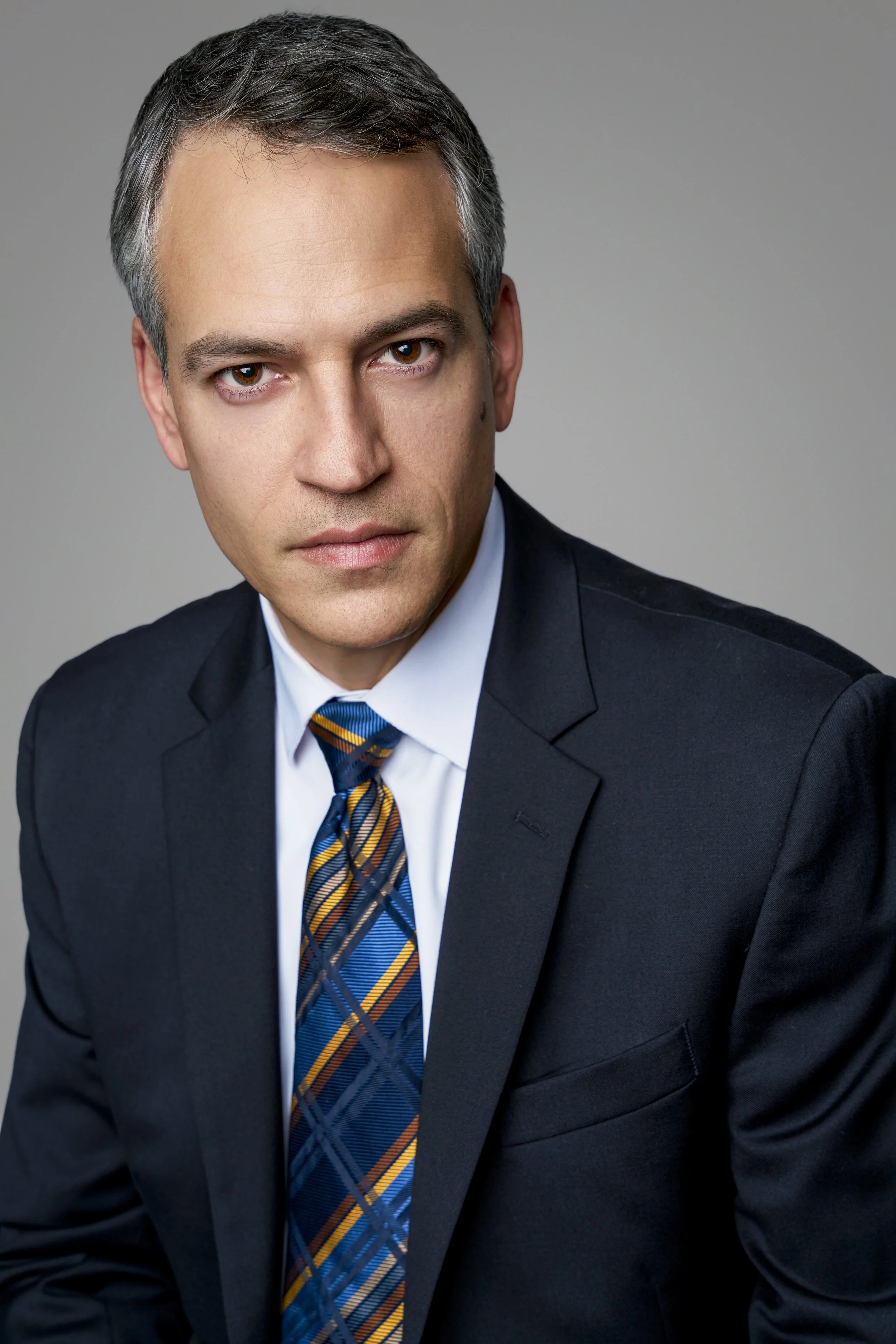 A man in a black suit, white shirt, and a colorful striped tie, looking at the camera with a serious expression, against a plain gray background.
