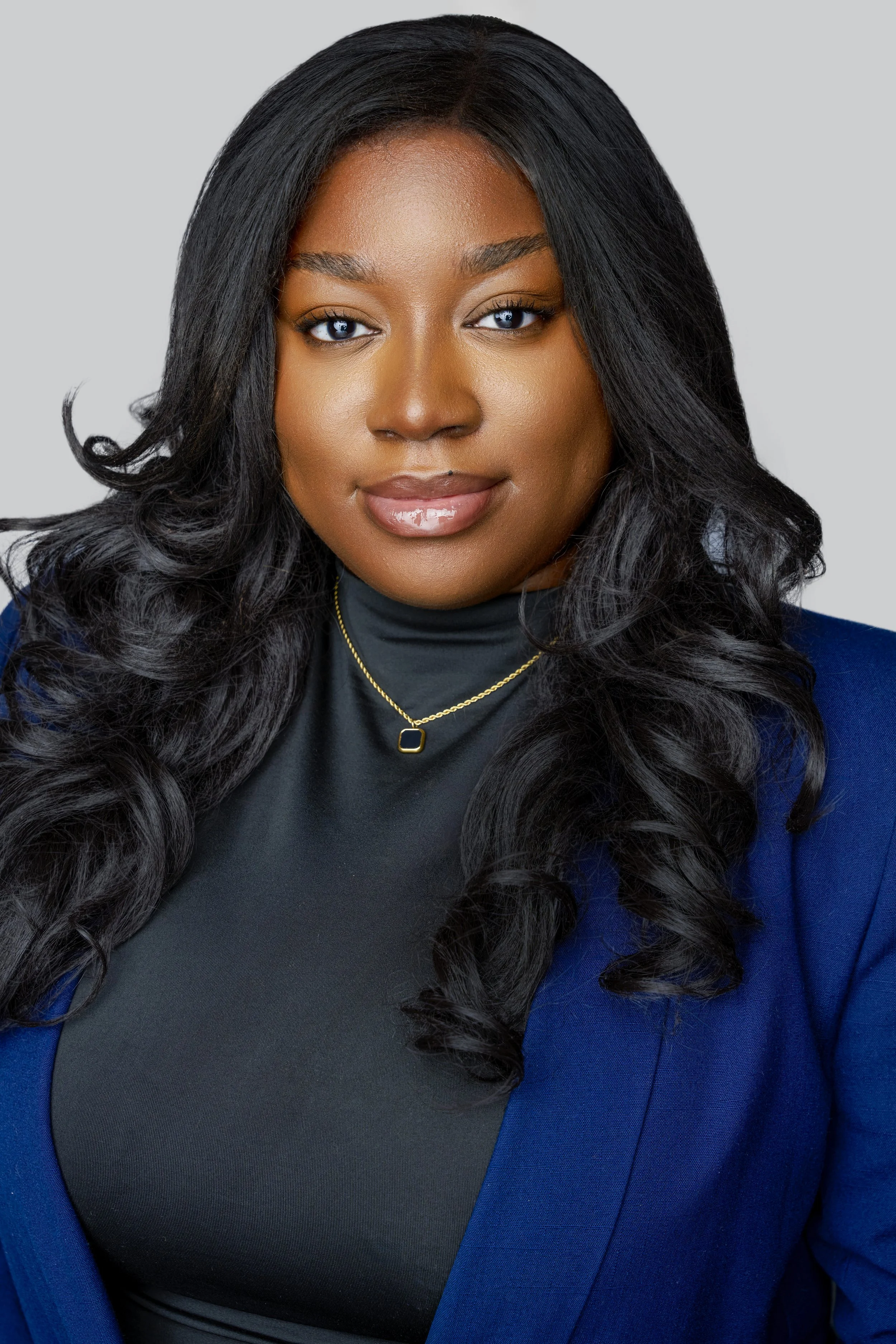 A portrait of a woman with long, black, curly hair wearing a black turtleneck, a blue blazer, and a gold necklace, against a light gray background.