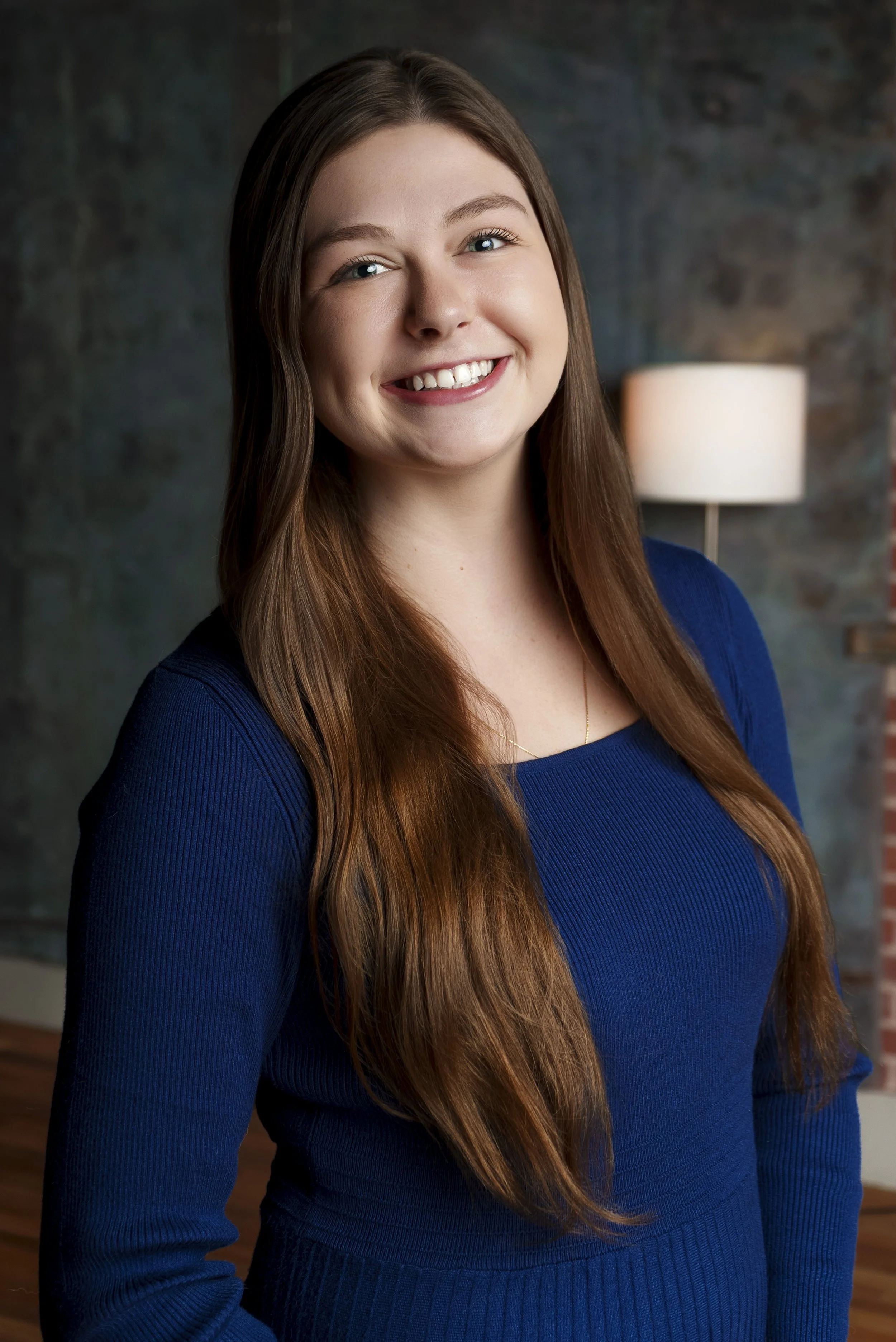 A young woman with long brown hair wears a blue top and smiles at the camera, standing in front of a textured dark background and a lamp.