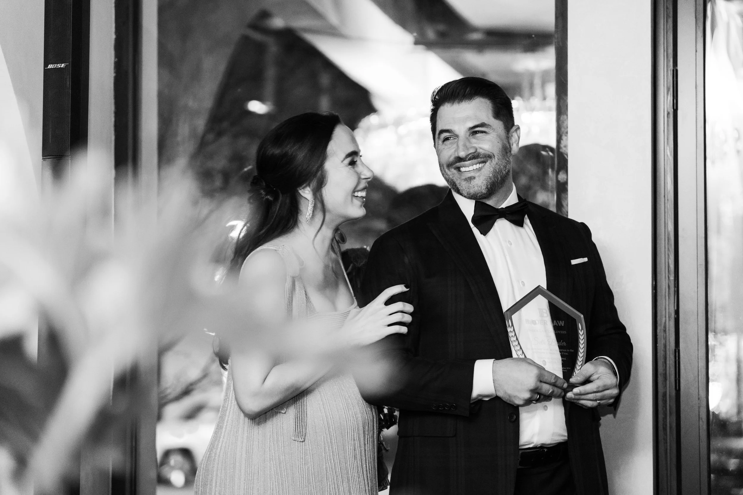 A black and white photo of a woman and man dressed in formal attire, smiling and interacting at an event. The woman is touching the man's arm, and he is holding a framed award or certificate.