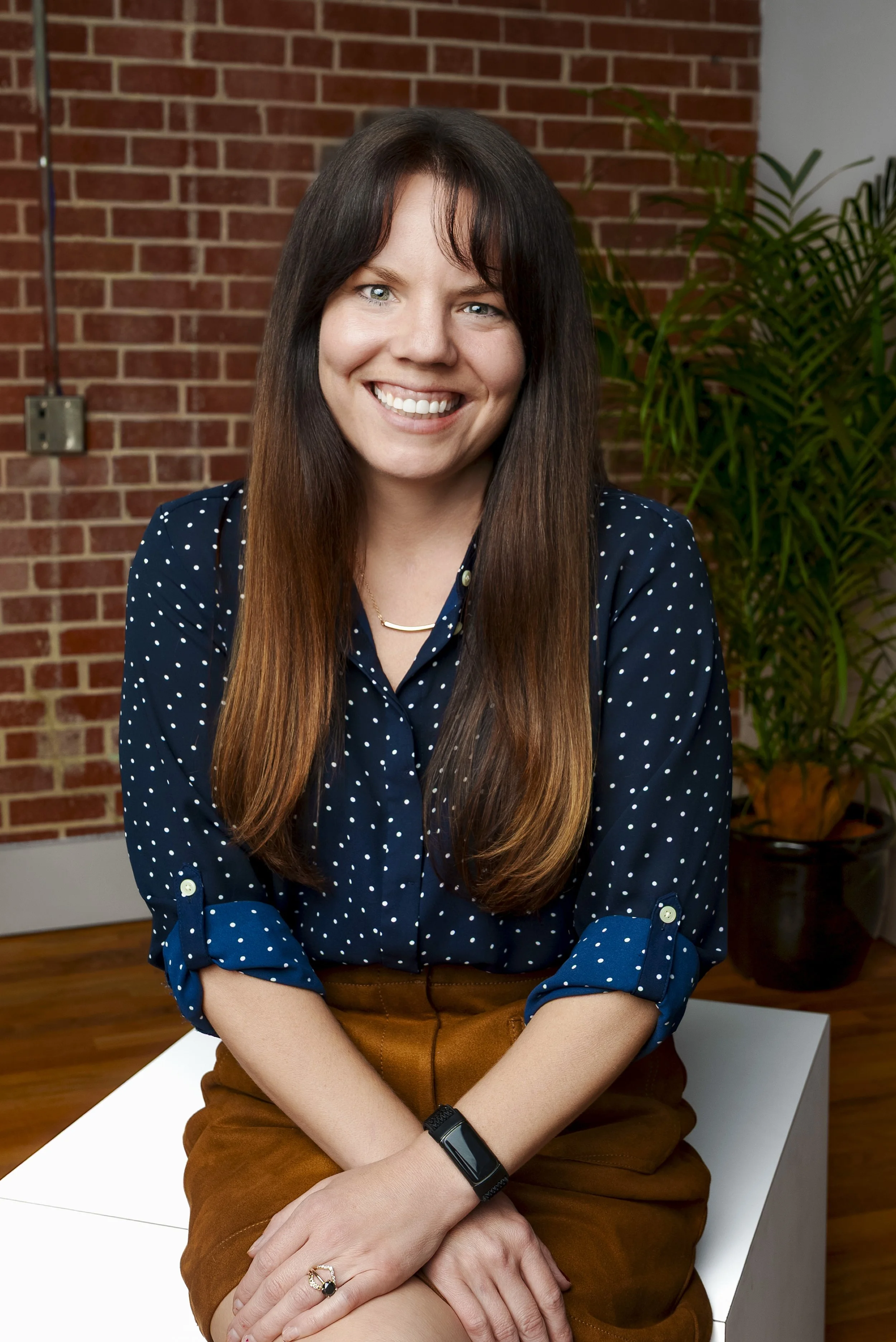 A woman with long brown hair and blue eyes smiling, wearing a navy blue polka dot blouse, sitting on a white block in front of a brick wall and green potted plants.