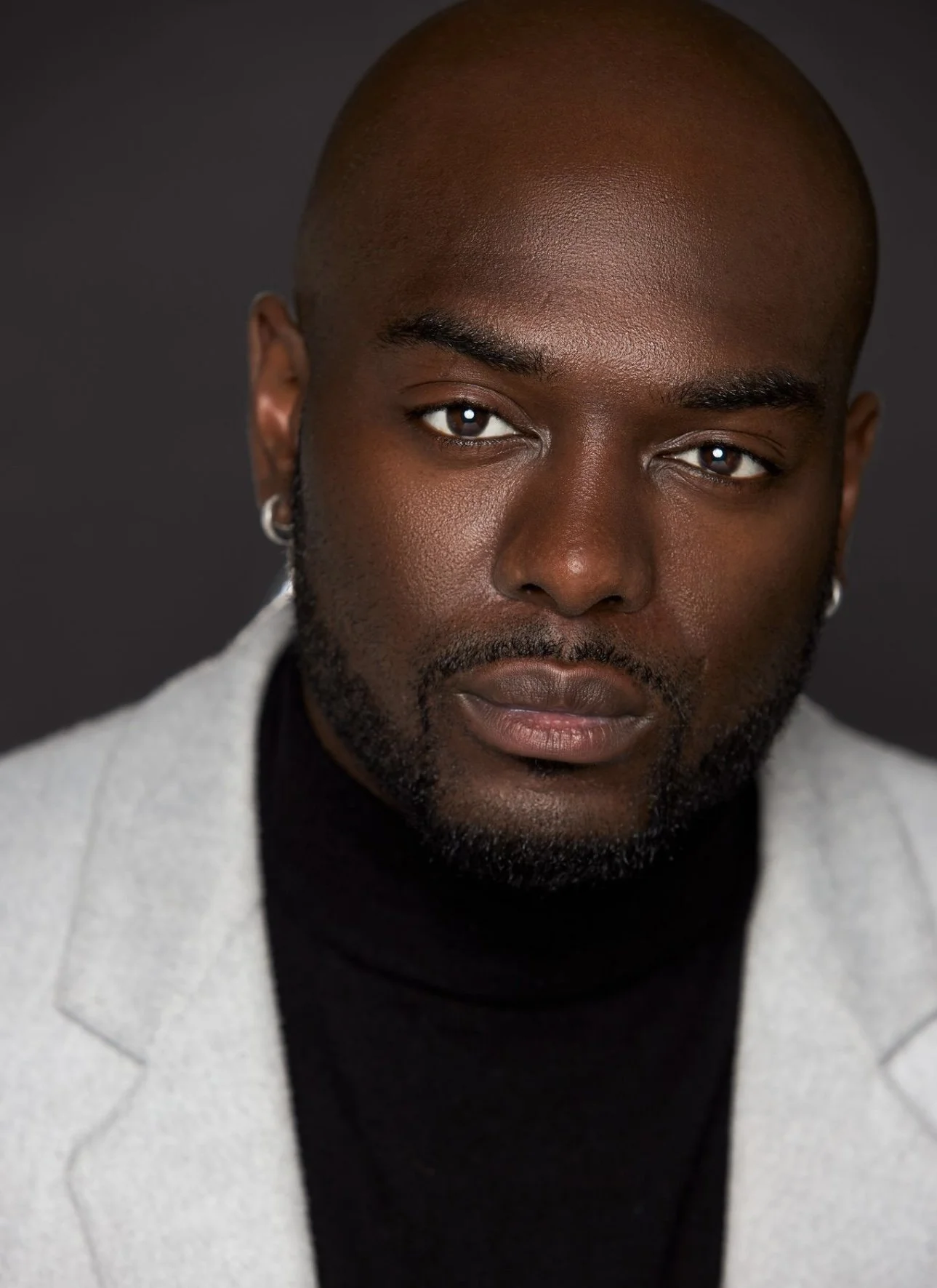 A close-up portrait of a Black man with a shaved head, wearing hoop earrings, a black turtleneck, and a light gray blazer, looking directly at the camera with a neutral expression against a dark background.