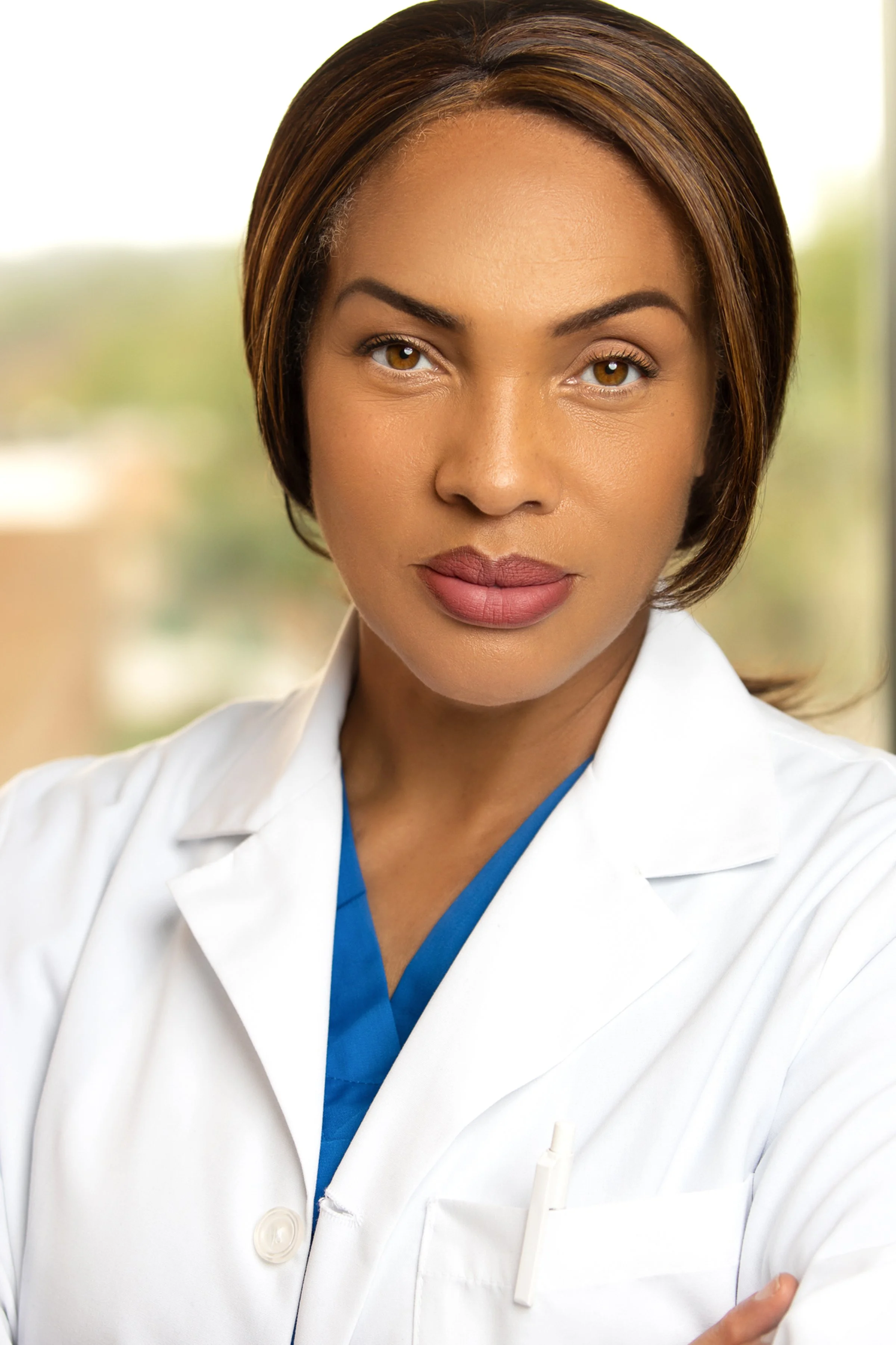 A female healthcare professional wearing a white lab coat and blue scrubs, looking confidently at the camera, with a blurred window scene in the background.