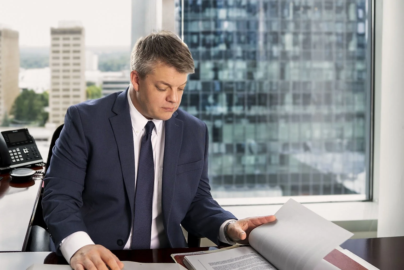 A man in a business suit sitting at a desk with a large window behind him, reading documents.