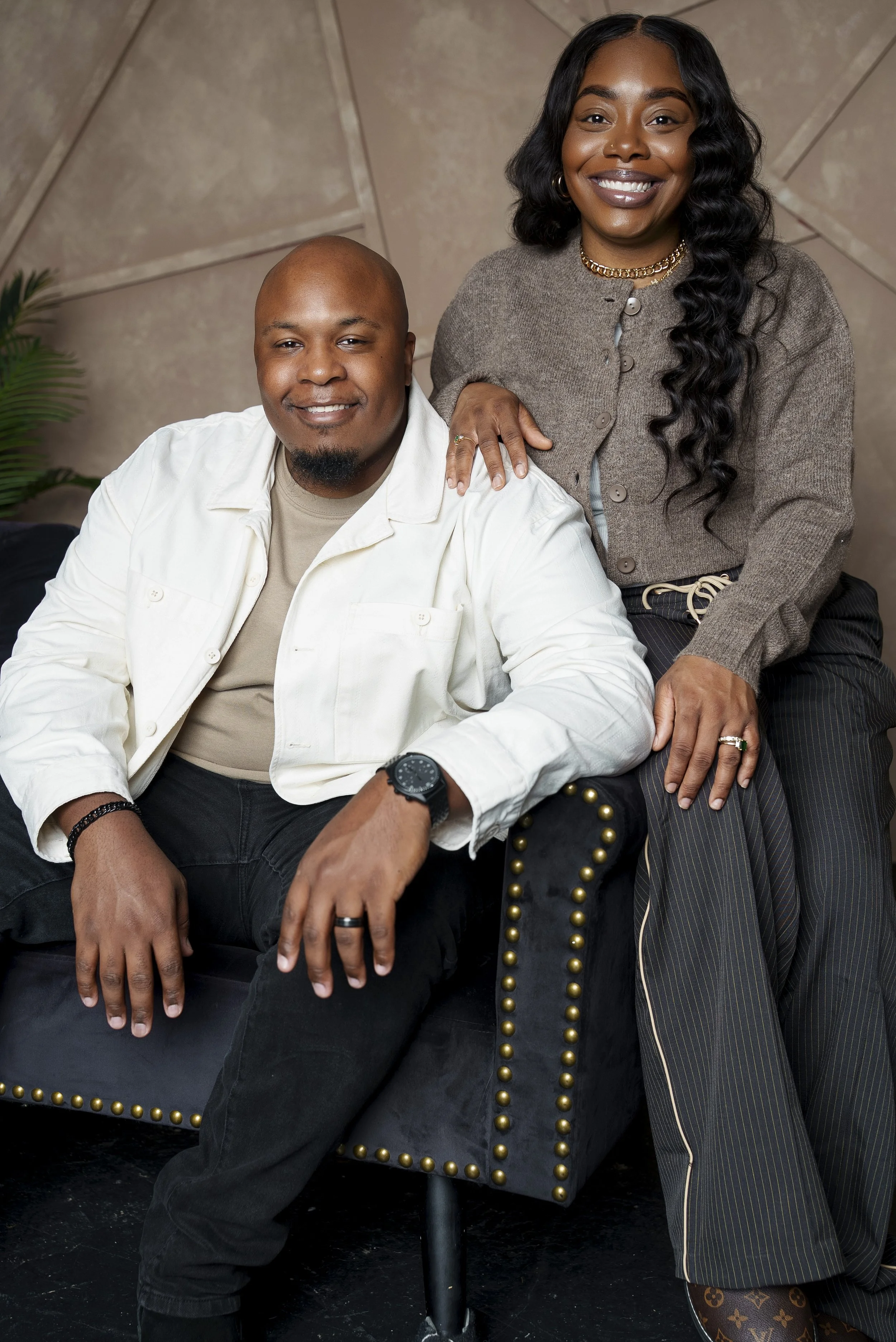 A smiling man and woman sitting closely together on black furniture, with the woman resting her hand on the man's shoulder, in a cozy indoor setting.