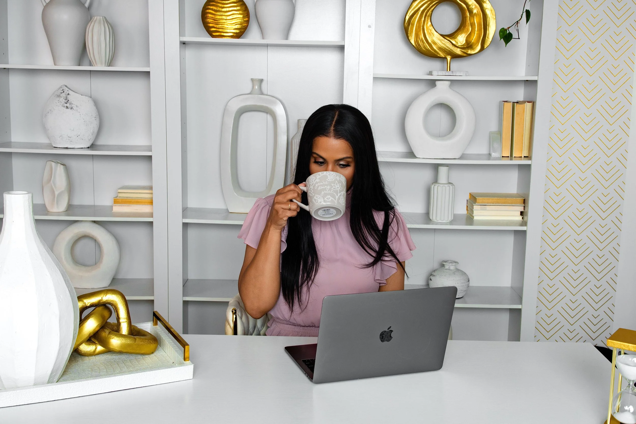 A woman with long black hair sitting at a white desk, drinking from a floral-patterned mug, with a silver MacBook laptop open in front of her. Behind her is a white bookshelf decorated with various white and gold vases and books, set against a wall w