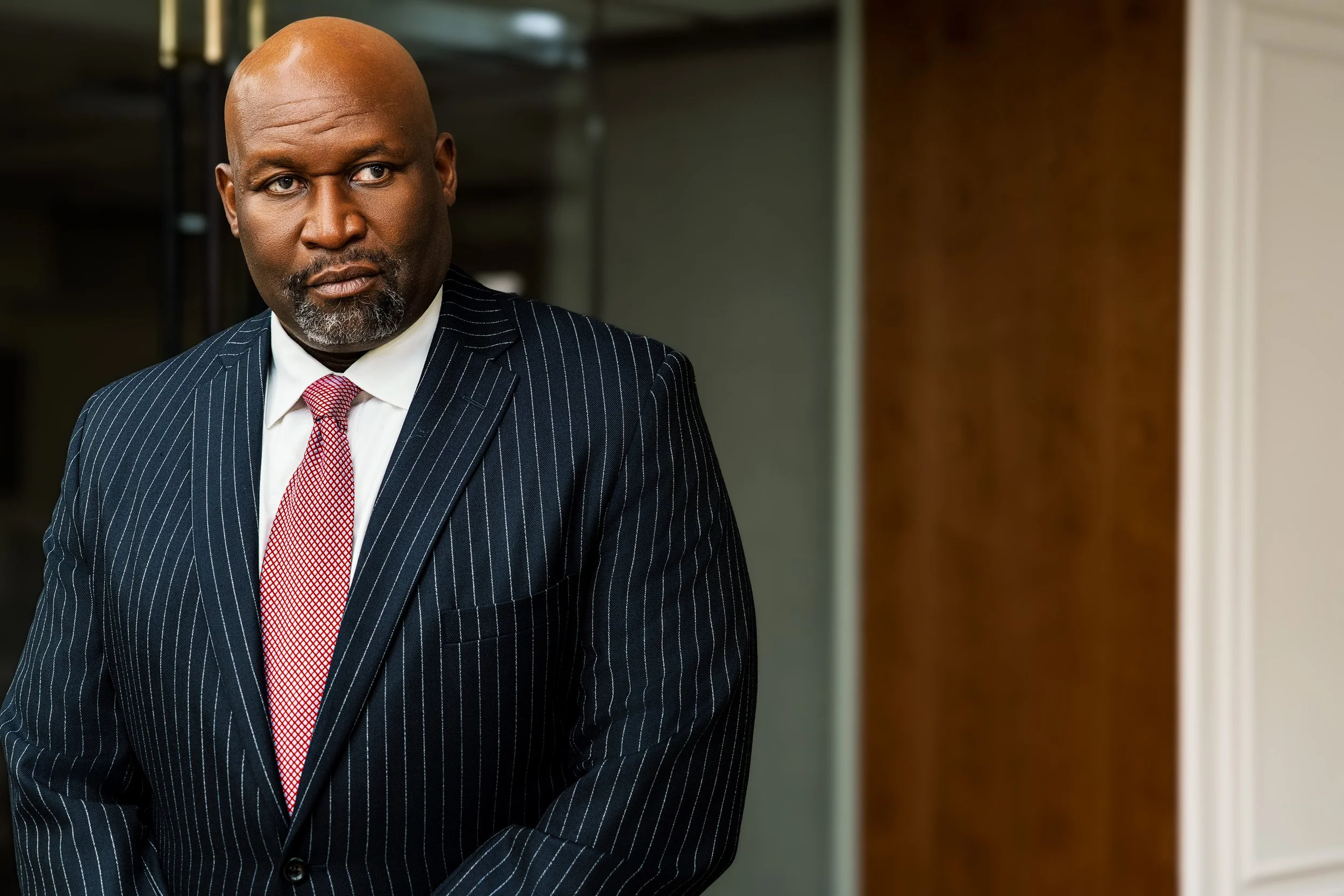 A middle-aged man with dark skin, bald head, and gray beard, wearing a dark pinstripe suit and a red tie, standing indoors near a wooden wall and a door frame.