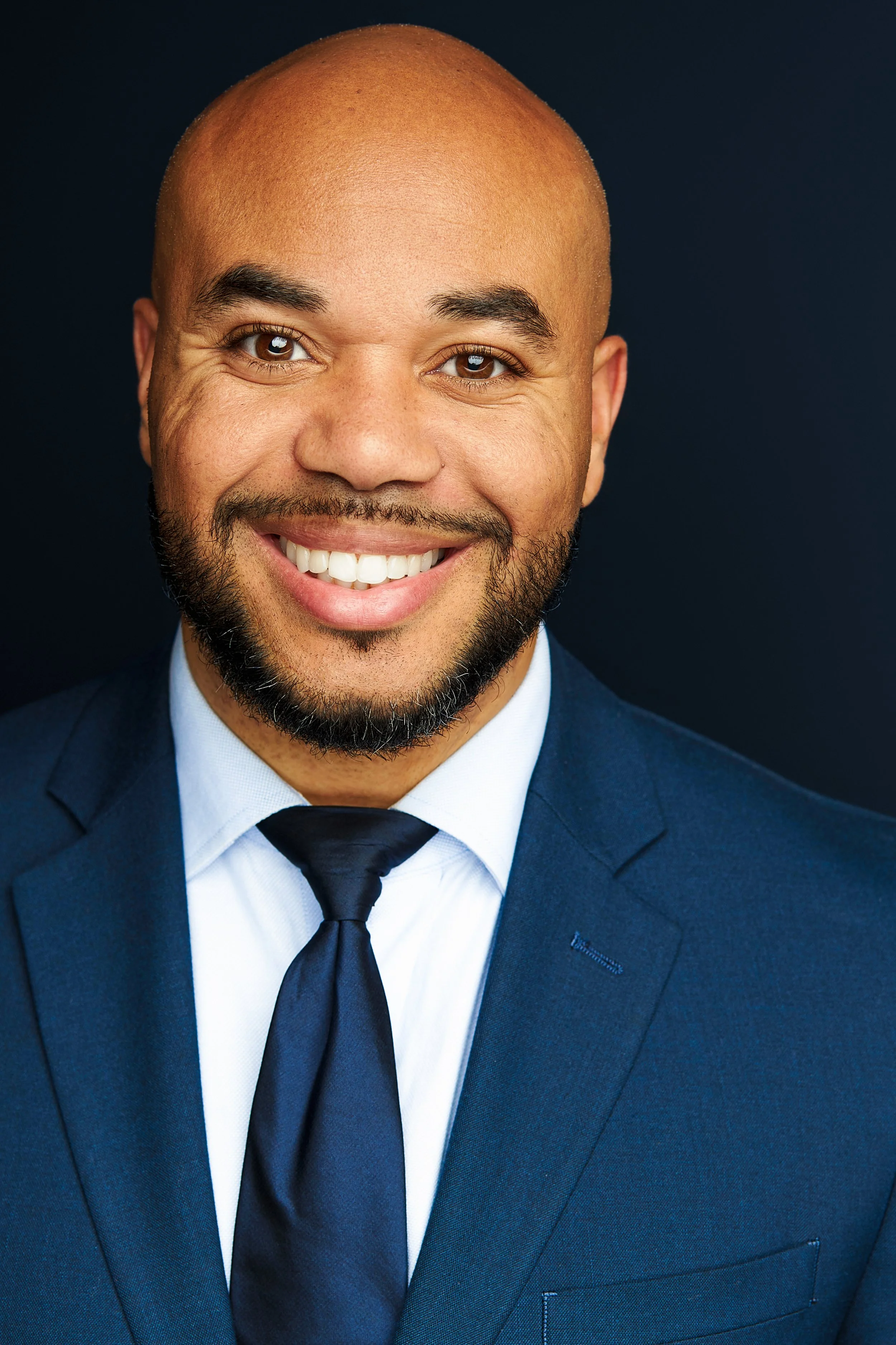 Professional headshot of a smiling man with a beard, dressed in a navy blue suit, white shirt, and black tie, against a dark background.