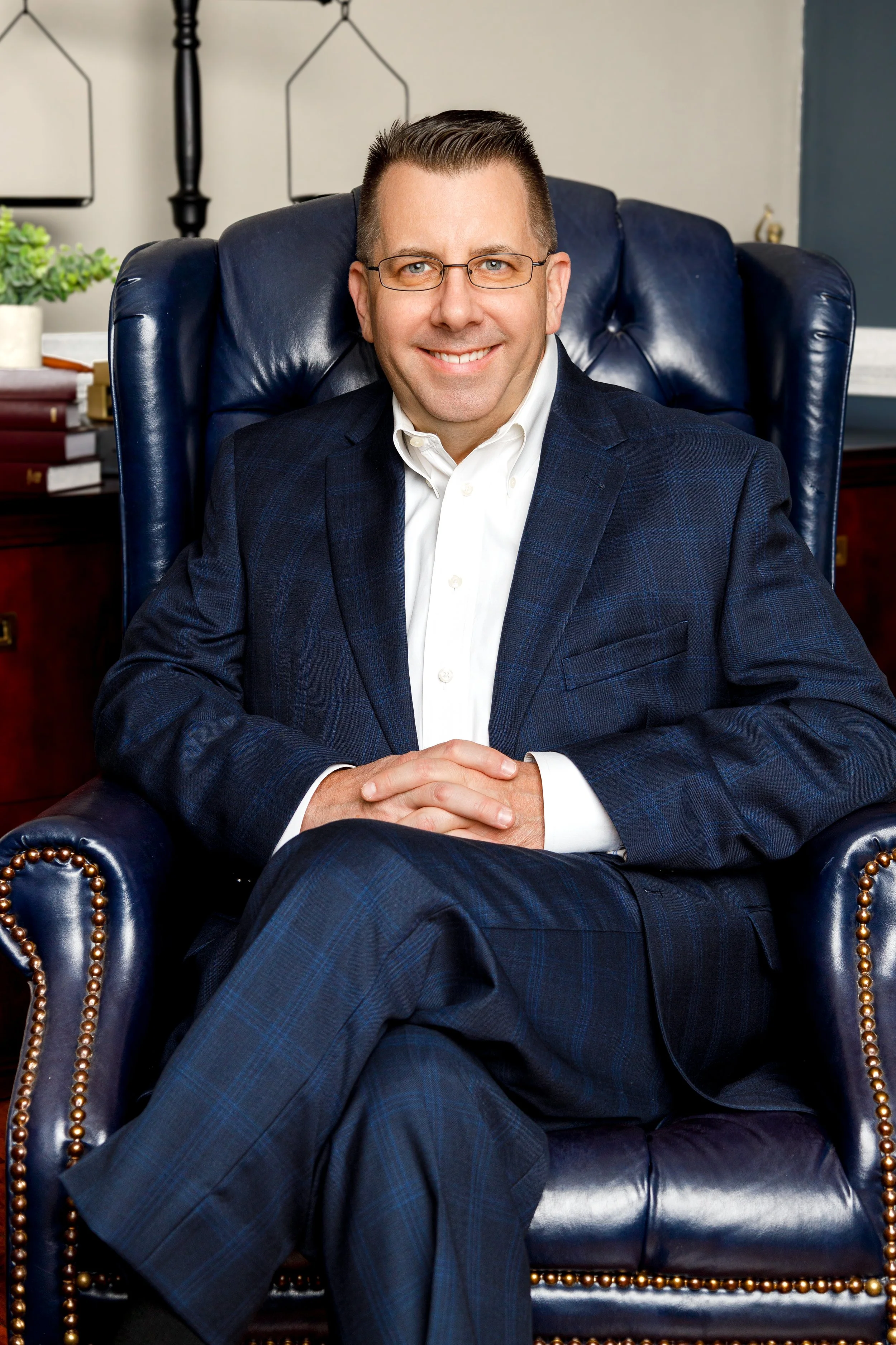 A man in a navy blue plaid suit, white shirt, and glasses sitting confidently in a large, dark leather armchair in an office setting with a desk, books, plant, and wall décor in the background.