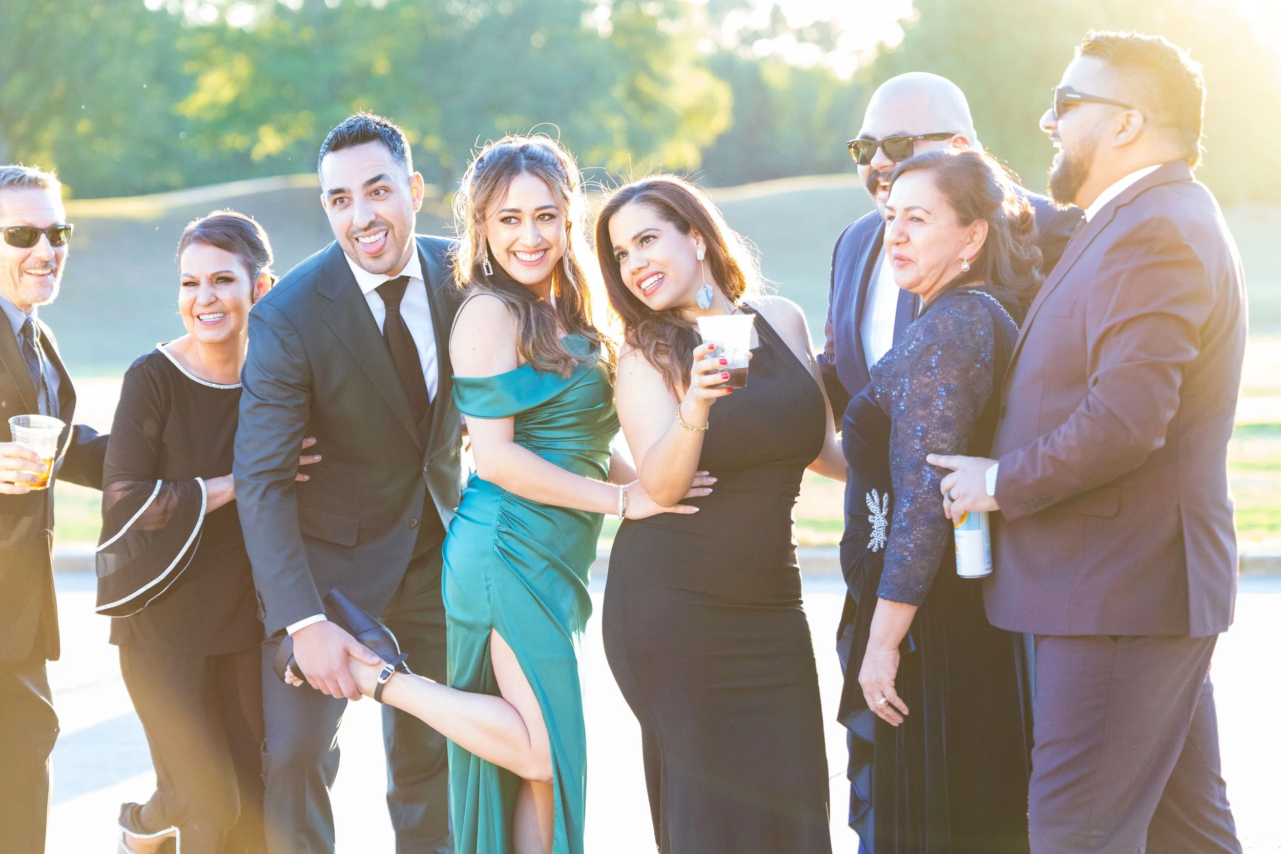 A group of nine adults in formal attire enjoying an outdoor gathering in sunny weather, with a park or golf course in the background.