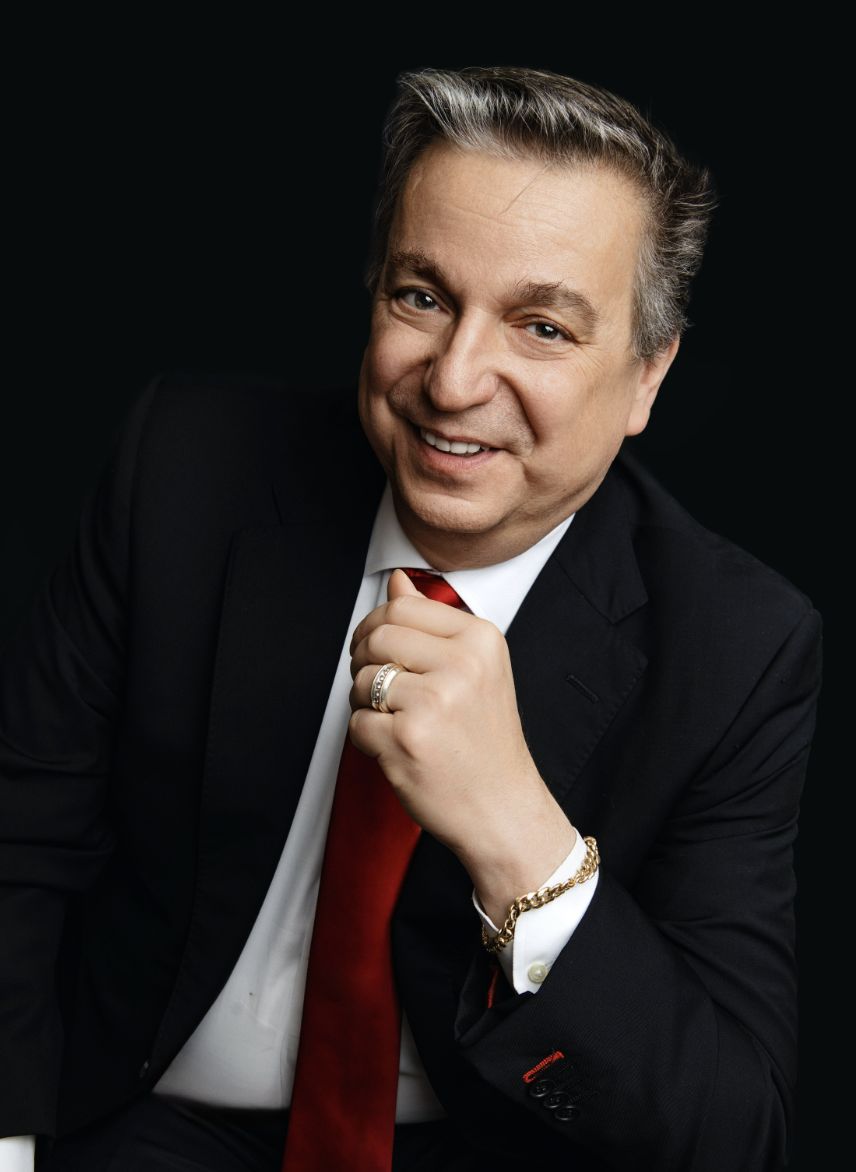 Portrait of a smiling middle-aged man in a black suit, white shirt, and red tie, with gold jewelry, against a dark background.