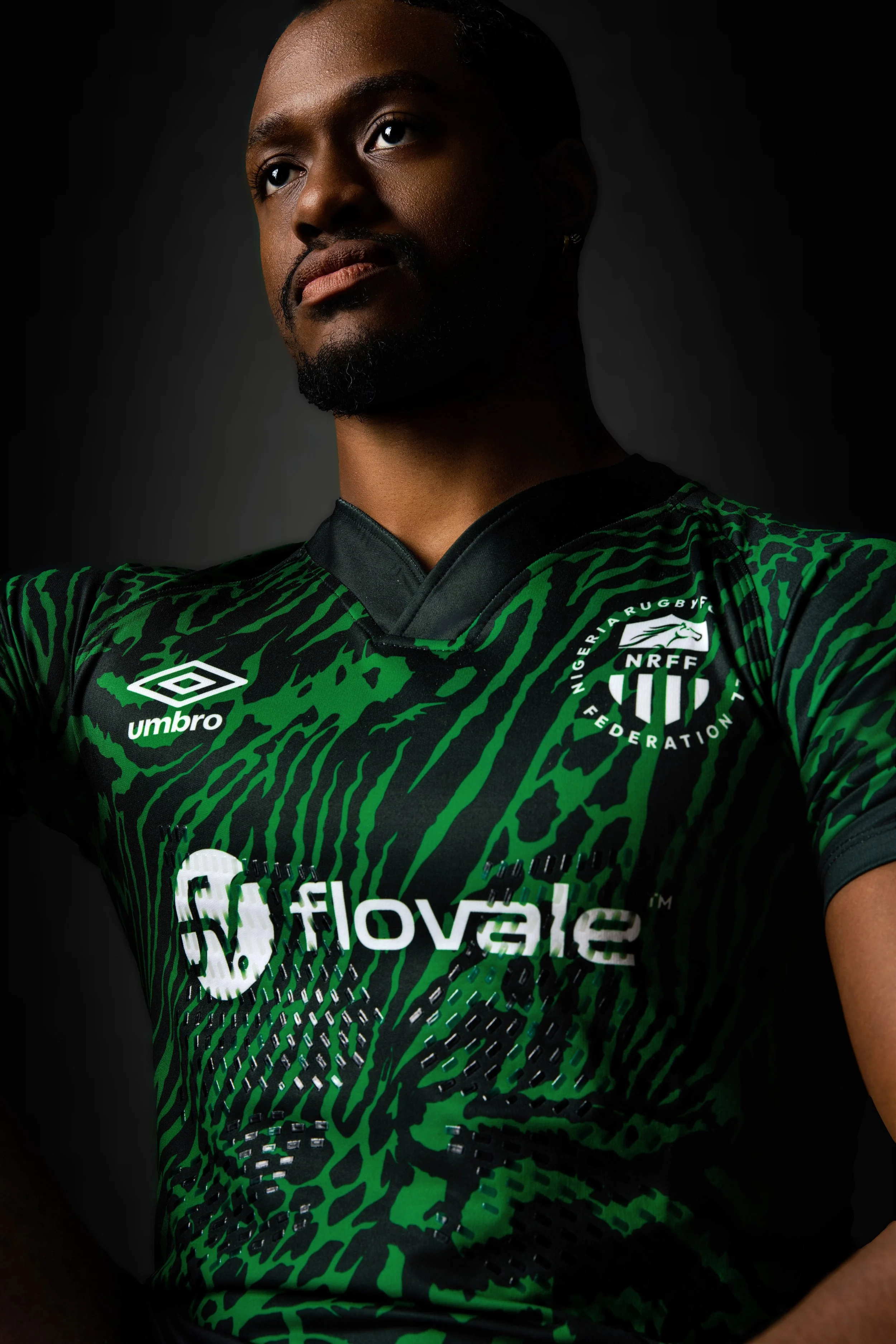 A man with dark skin, short hair, and a beard, wearing a black and green sports jersey with logos, looking upward.