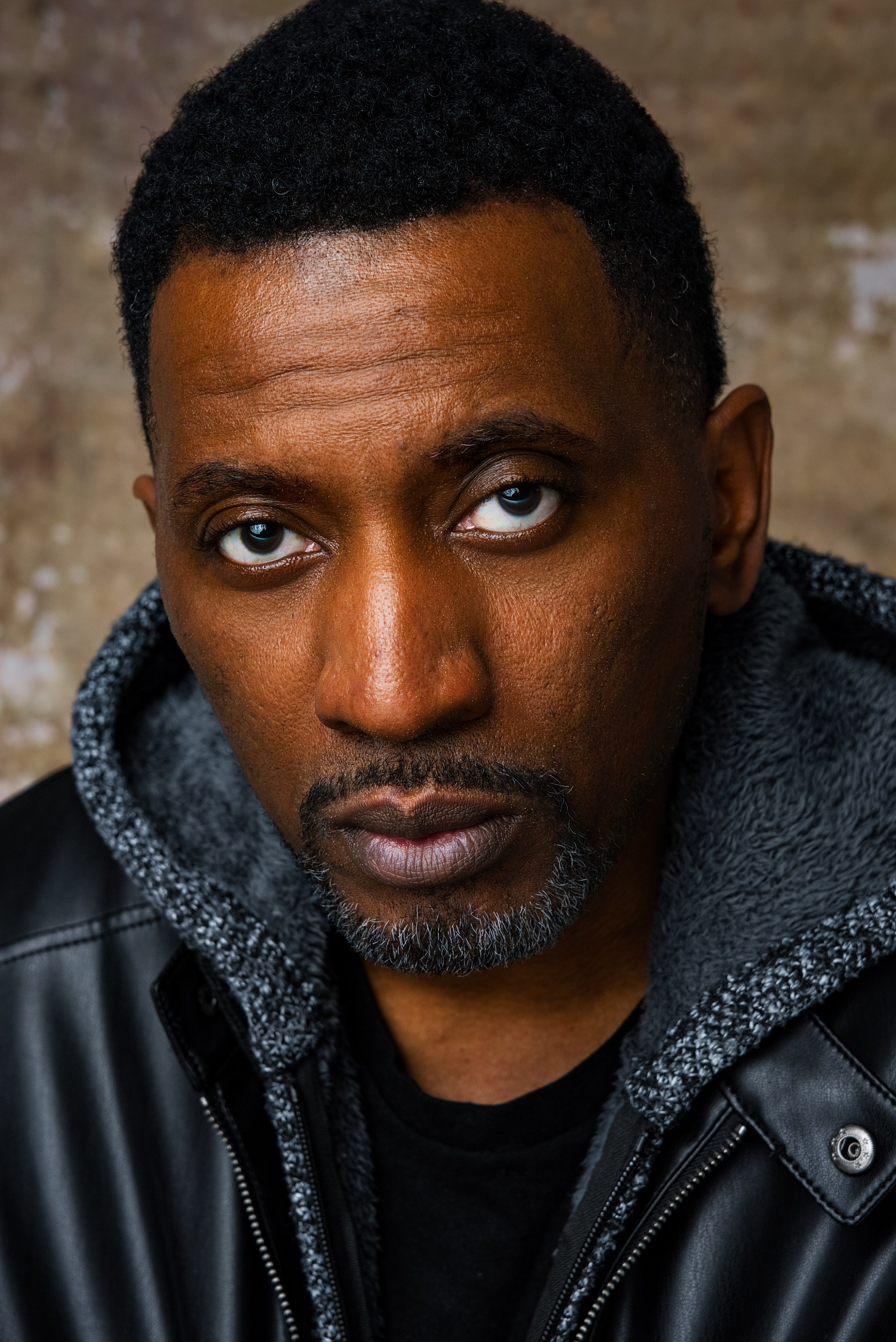 A close-up portrait of a man with dark skin, short curly black hair, and light blue eyes, wearing a black leather jacket with a gray hoodie underneath, against a blurred brick wall background.
