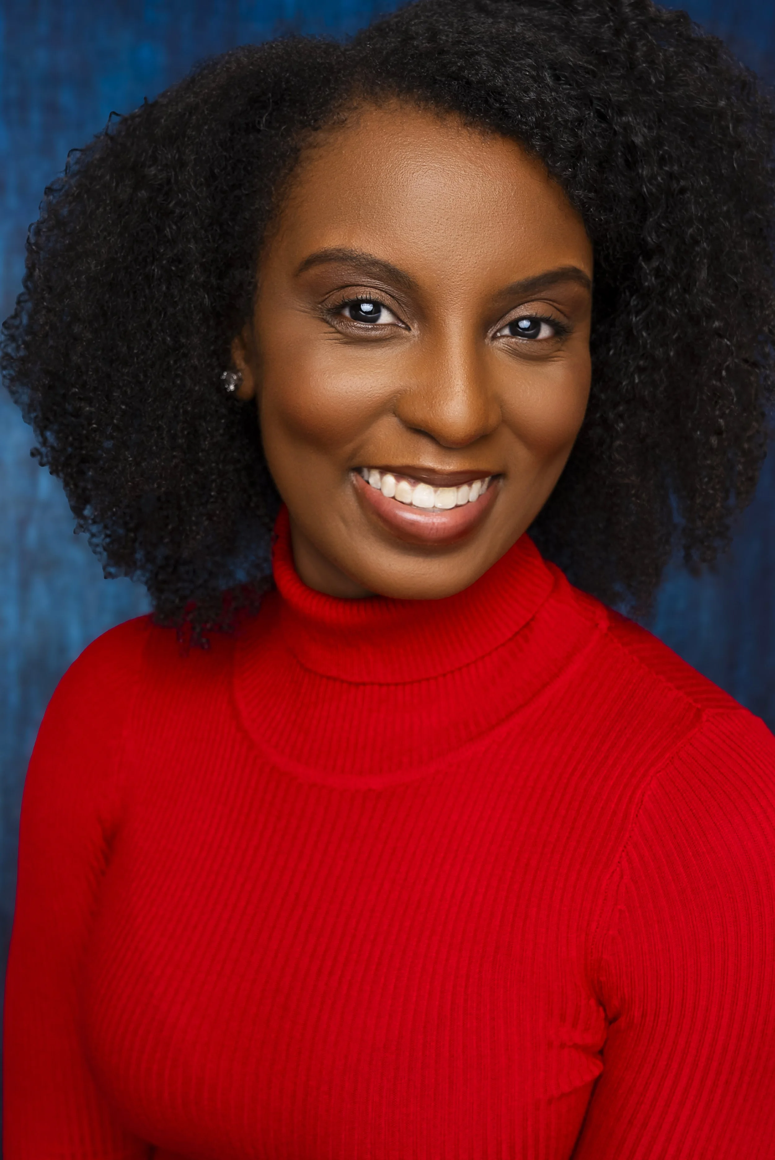 A woman with curly black hair, wearing a red turtleneck sweater, smiling, with a blue background.