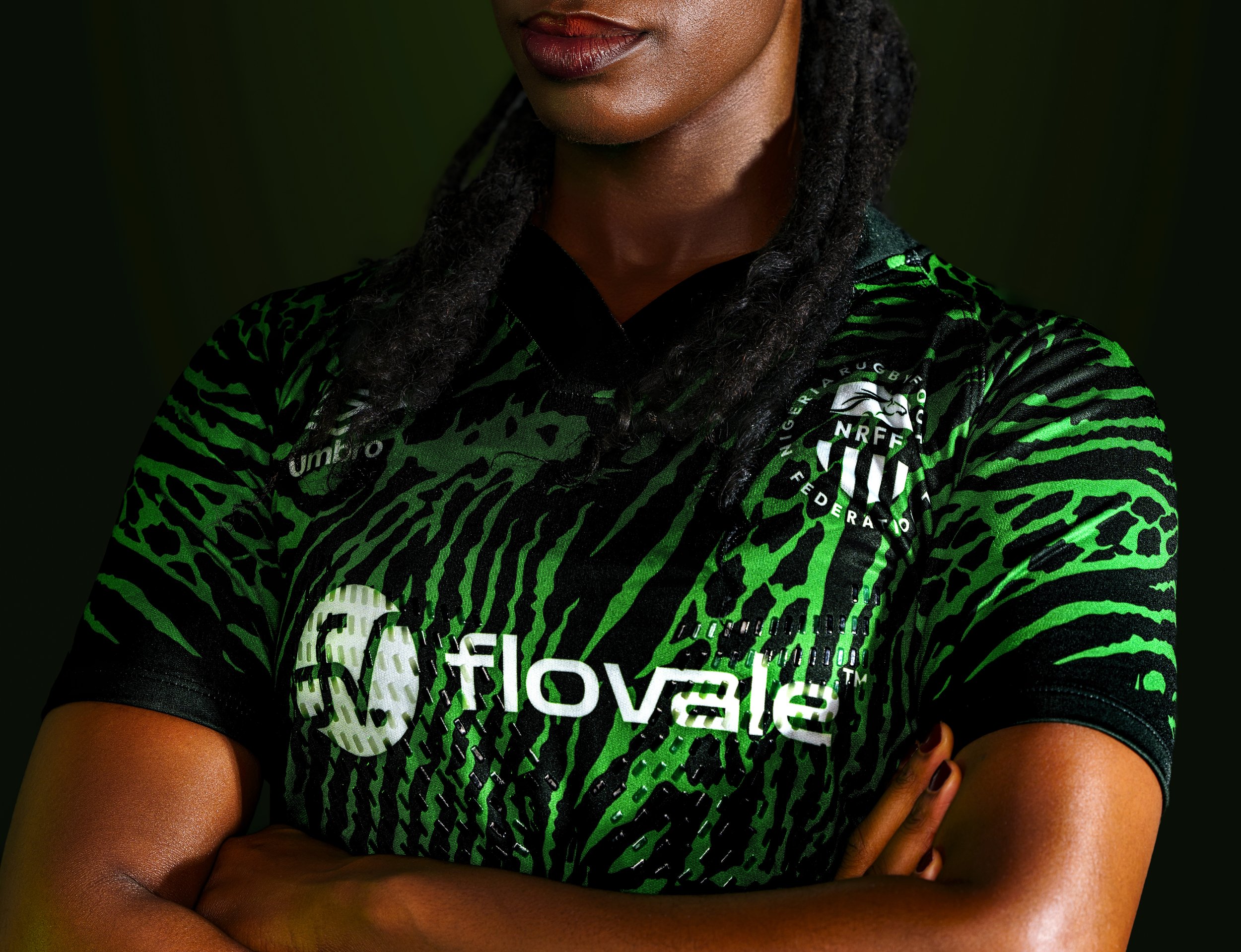A woman with dark skin and braided hair wearing a green and black sports jersey with logos and text, including 'NRFF Federation' and 'Umbro.' She has her arms crossed and is standing against a dark background.