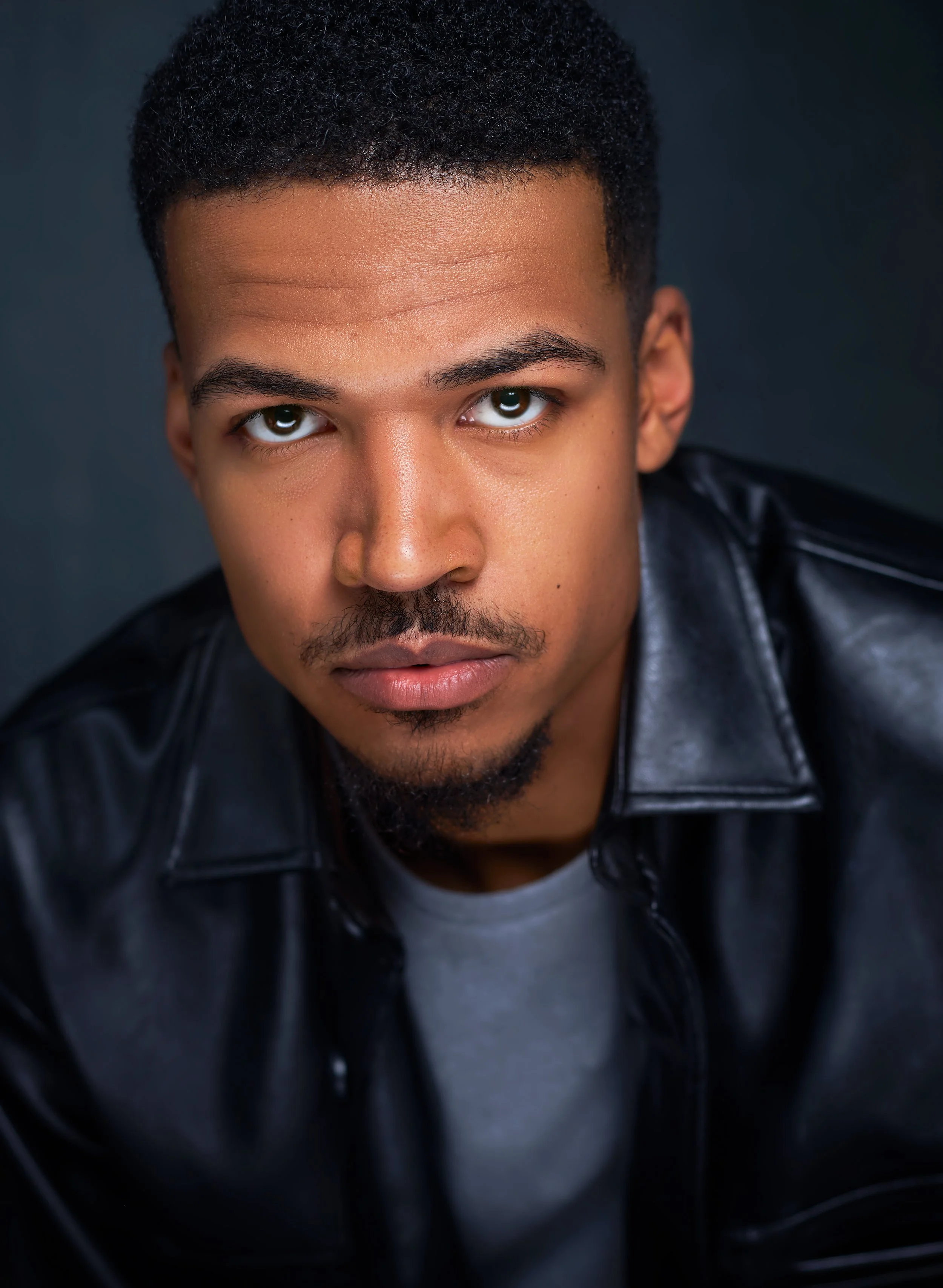 Close-up portrait of a young man with short curly black hair, brown eyes, and a thin mustache and goatee, wearing a black leather jacket.