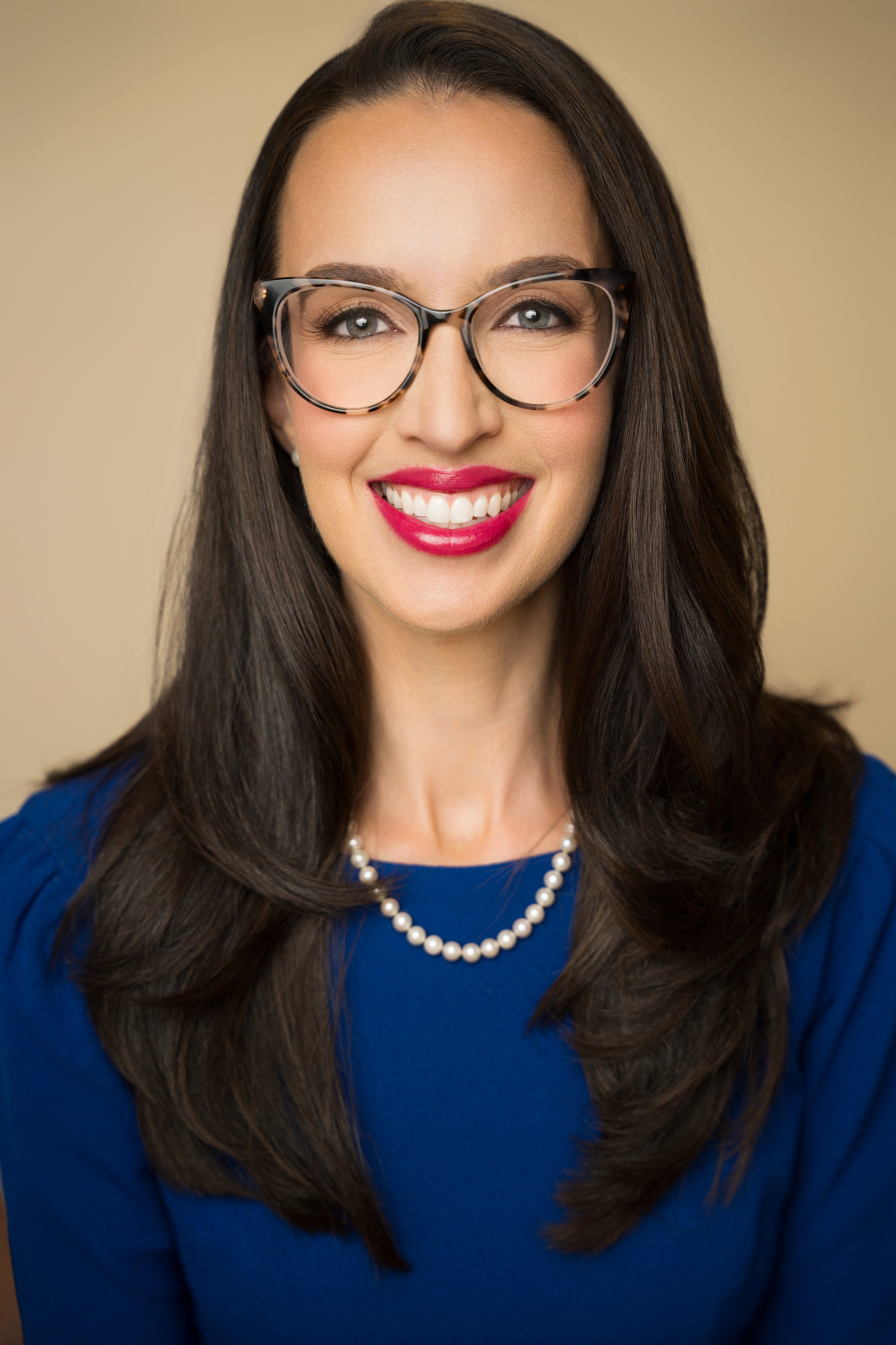 A woman with long dark hair wearing glasses, a pearl necklace, and a blue dress, smiling facing the camera.