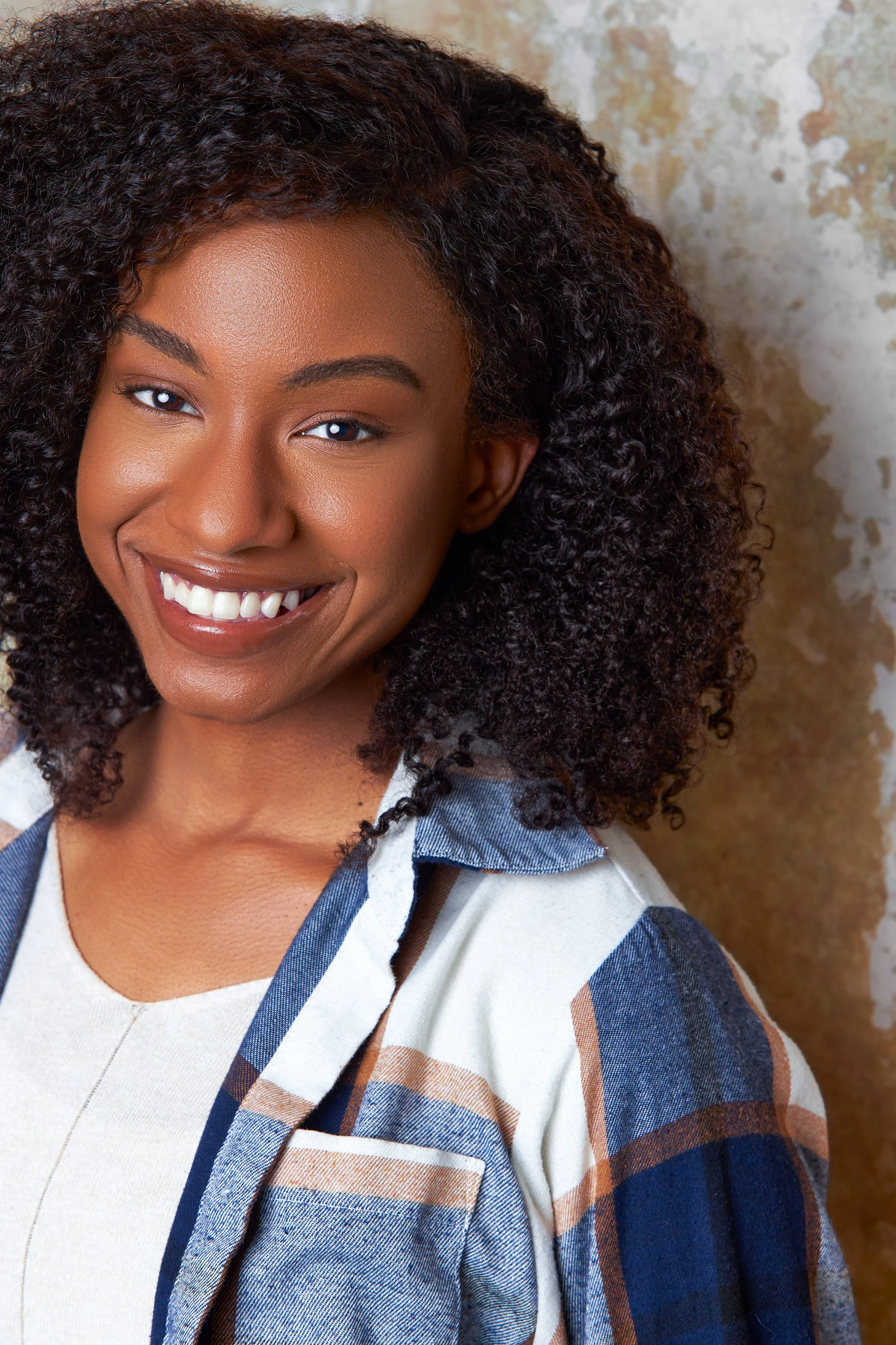Close-up of a smiling African American woman with curly hair, wearing a plaid shirt over a light top, standing against a textured wall.