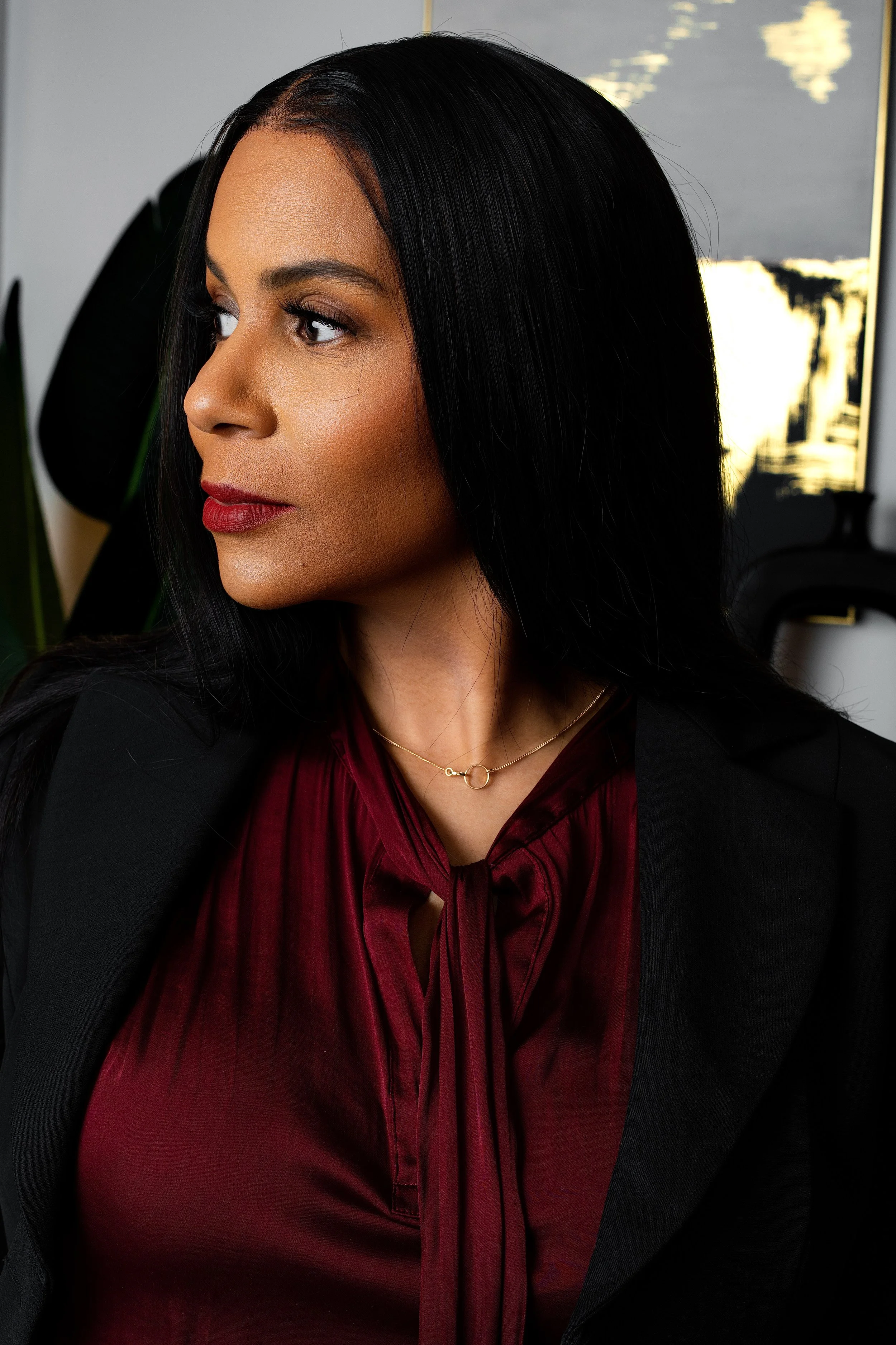 A woman with long black hair, wearing a black blazer and a dark red blouse, looking to her left in an indoor setting with a modern decor background.