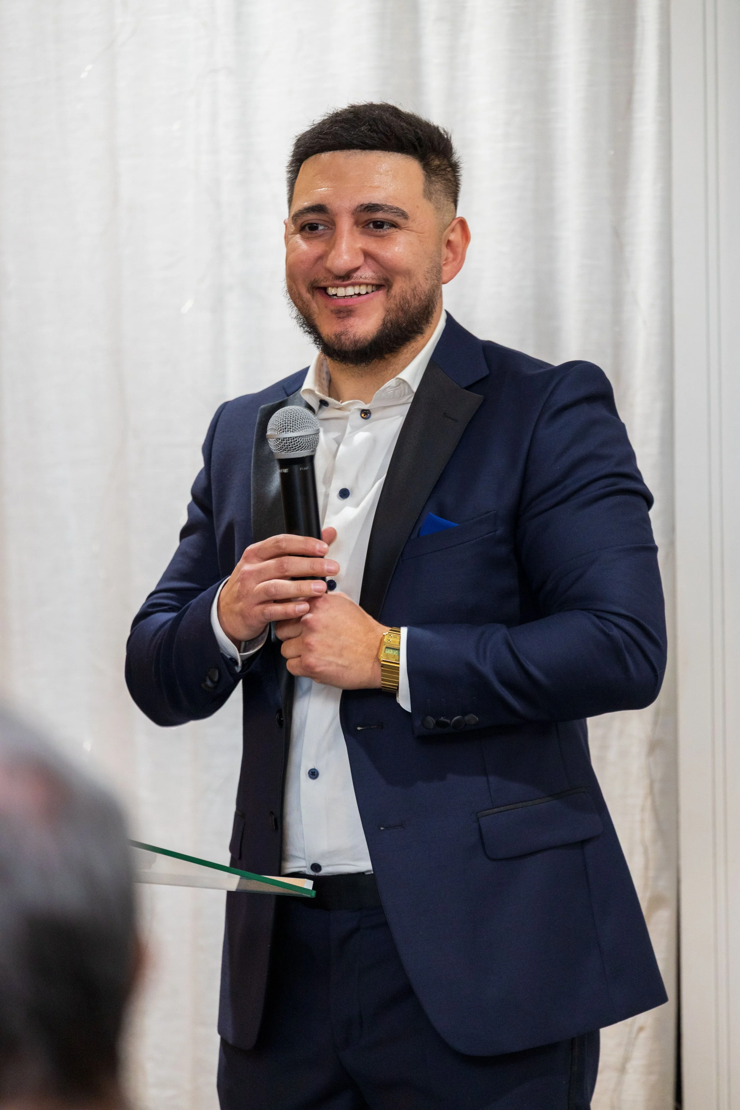 A man in a dark blue suit and white shirt smiling and holding a microphone, standing in front of a light-colored curtain.