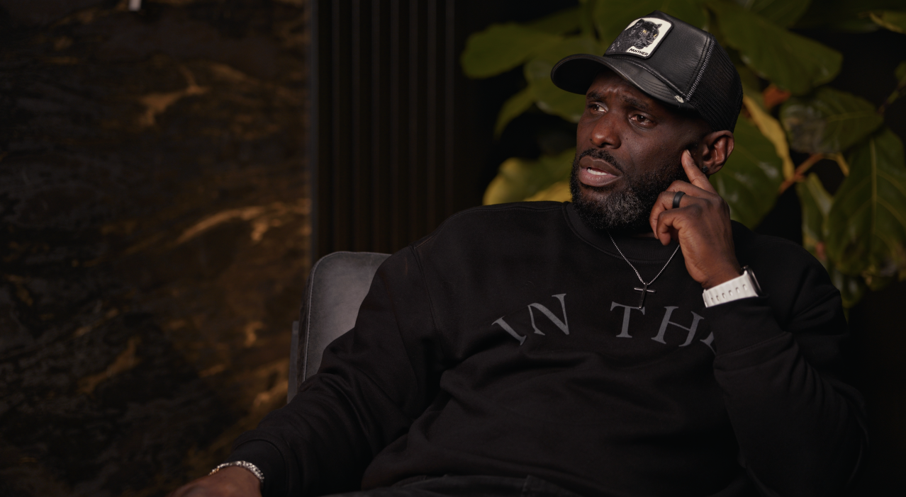 A man with dark skin, black beard, and mustache, wearing a black baseball cap, black sweater, silver chain, and white wristwatch, seated indoors near a large green plant, with a dark background and a textured wall.