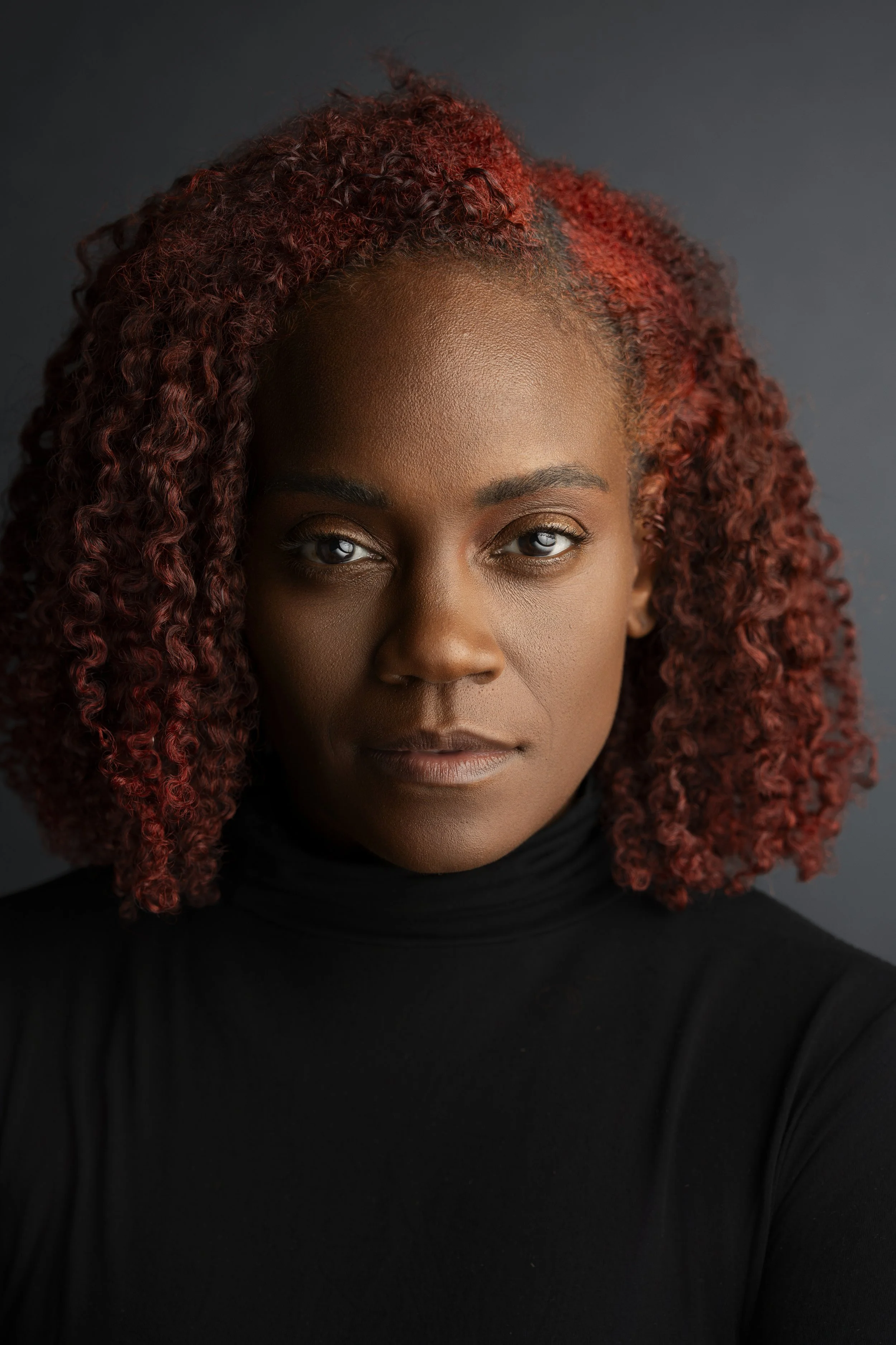 A woman with curly red hair wearing a black turtleneck poses against a dark background.