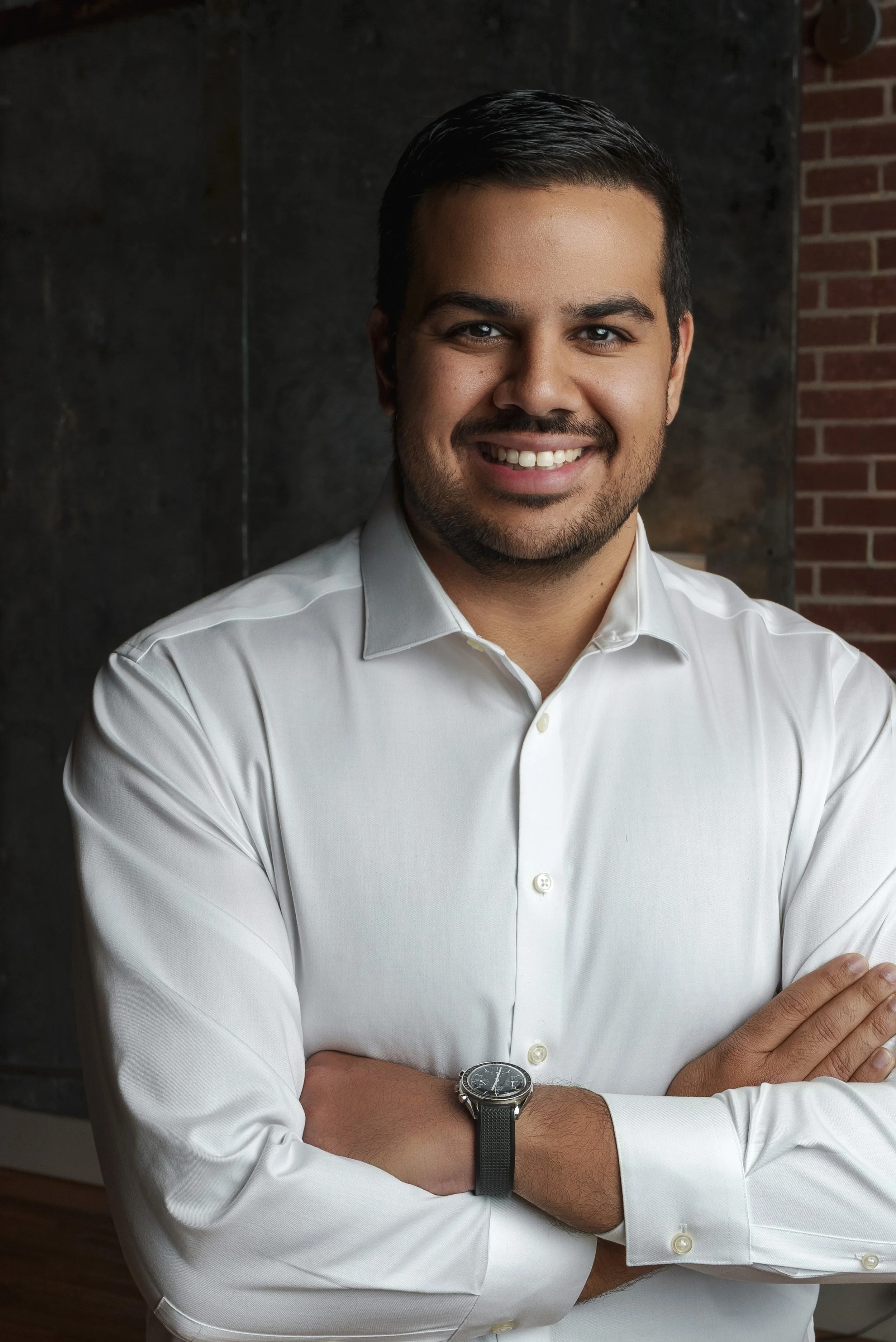 A smiling man in a white dress shirt with arms crossed, standing in front of a dark wall and brick background.
