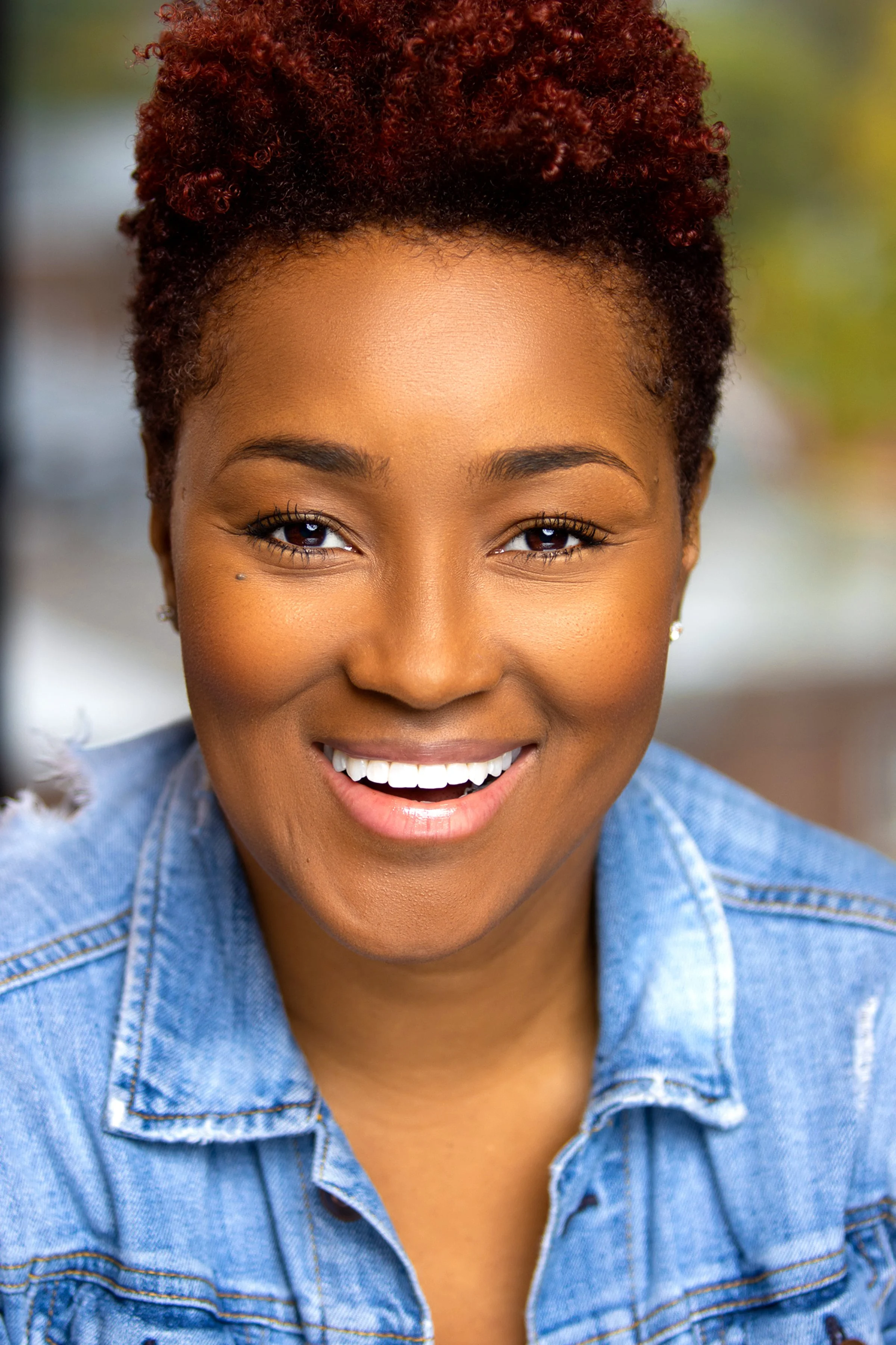 Close-up of a smiling woman with short, curly red hair, wearing a denim jacket and small stud earrings, outdoors with blurred background.