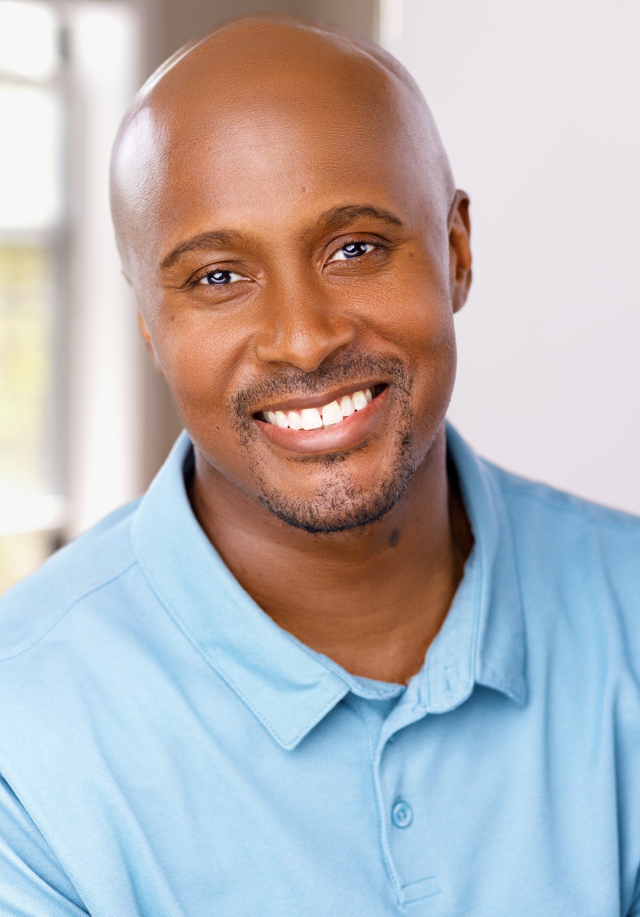 Close-up of a smiling man with a shaved head wearing a light blue collared shirt.