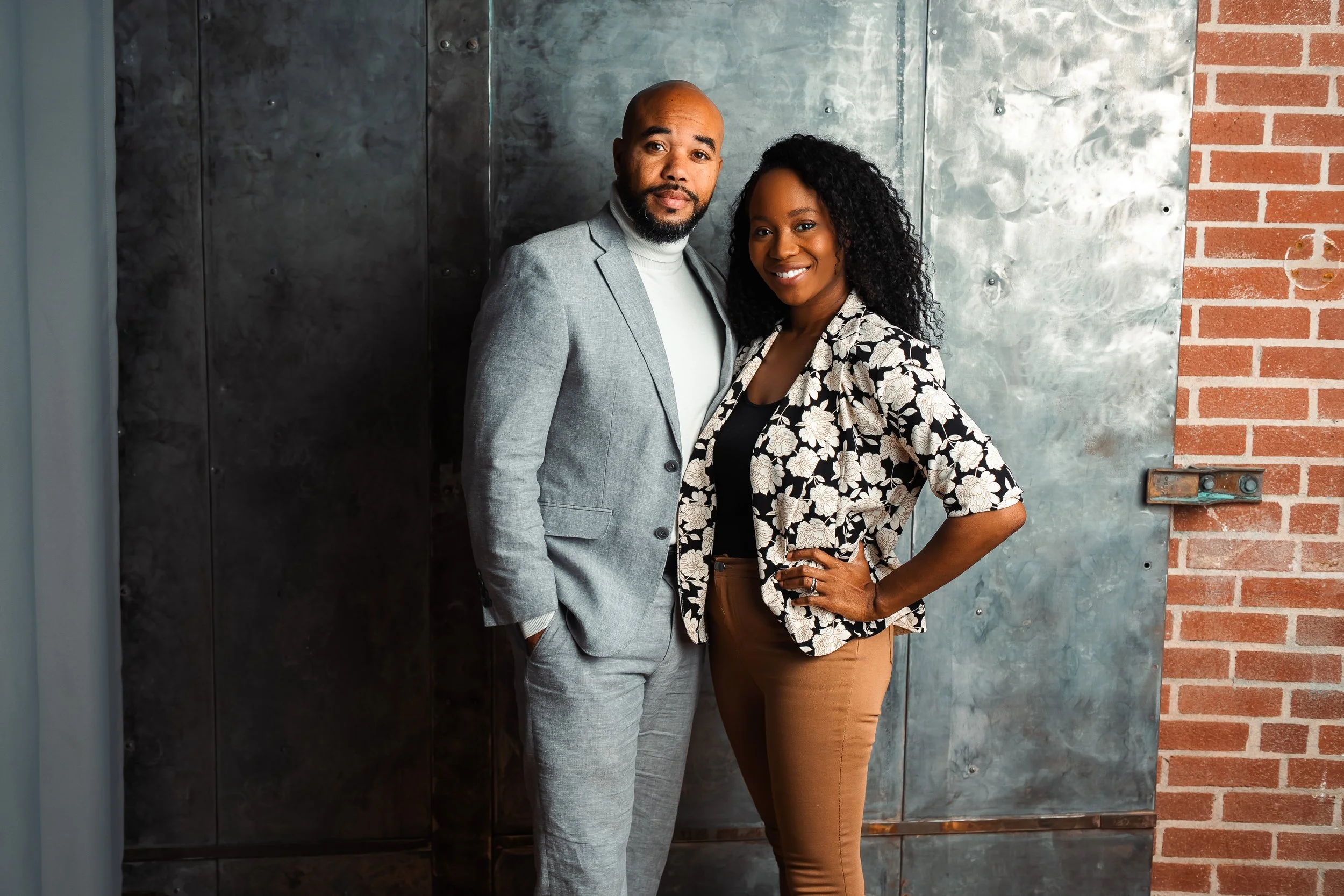 A man and woman standing together, smiling, in front of a industrial-style wall with metal and brick sections.