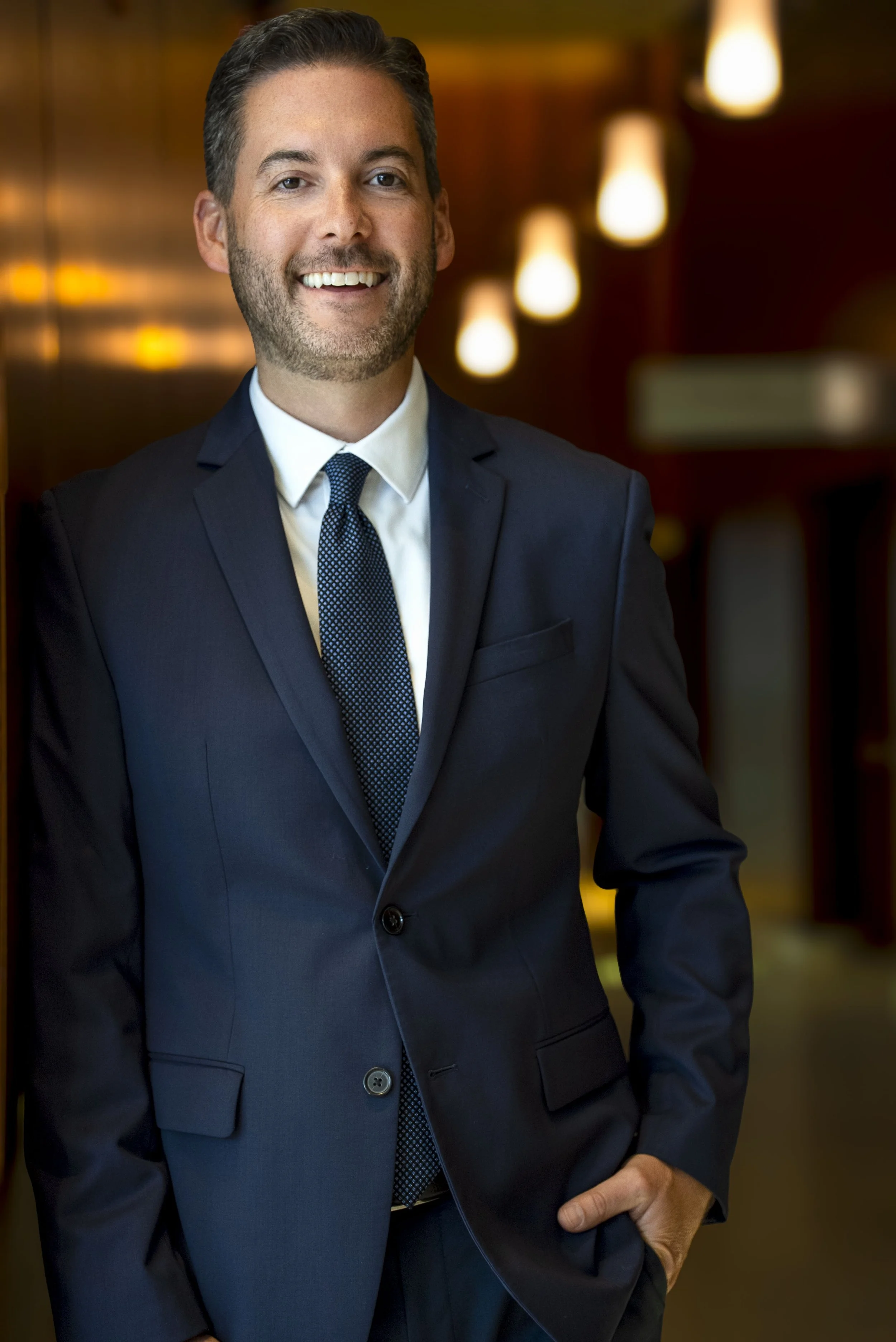 A man in a dark suit and tie smiling inside a warmly lit room.