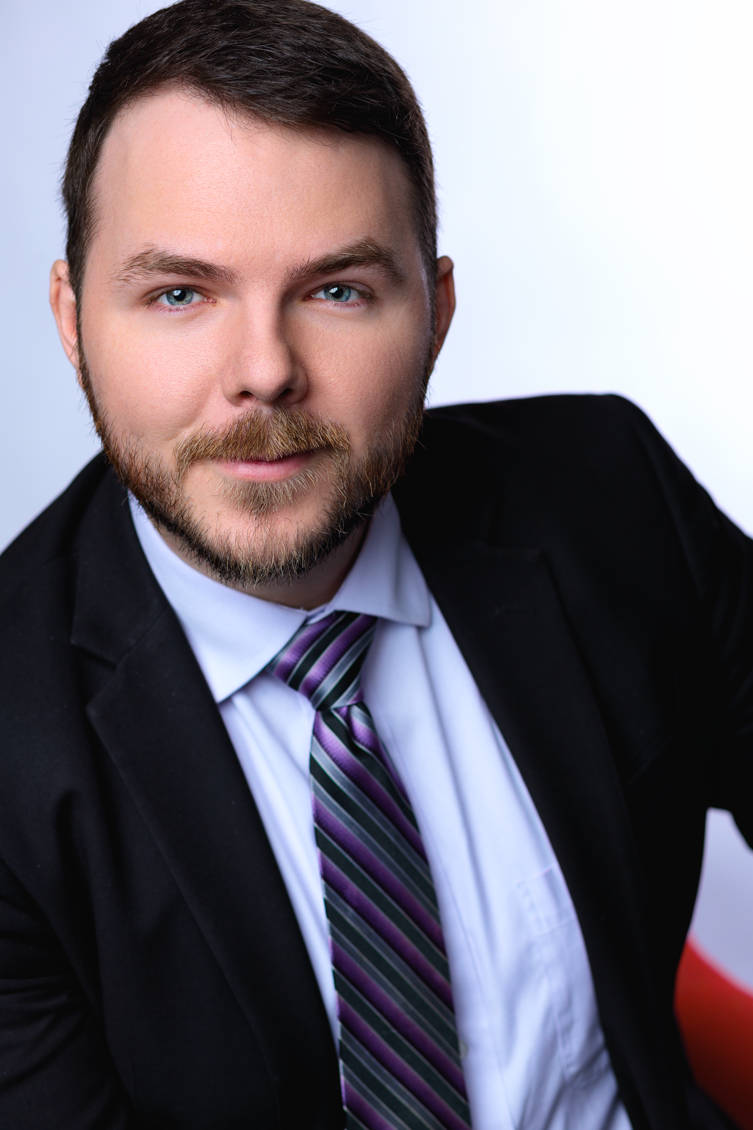 A young man with brown hair and a beard, dressed in a black suit, white shirt, and a purple striped tie, smiling at the camera.