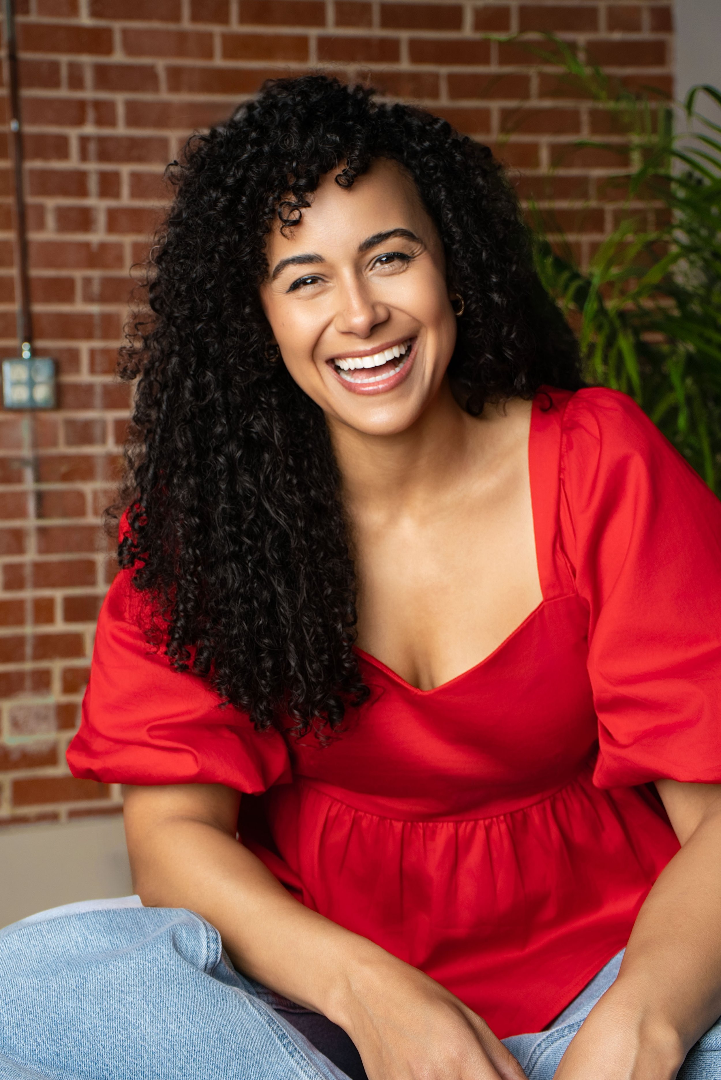 Smiling woman with curly hair wearing a red top sitting indoors in front of a brick wall and green plant.