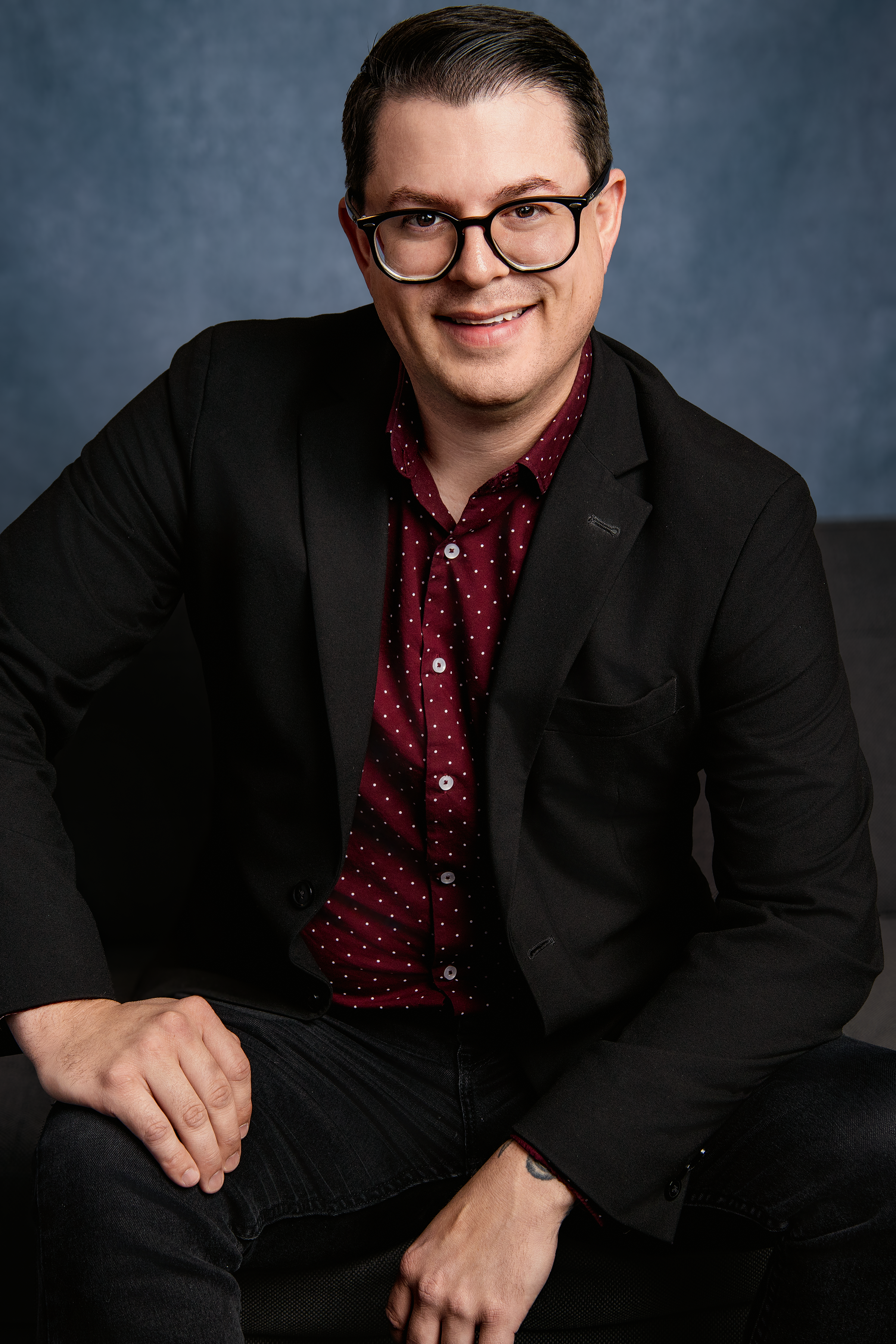 A man wearing glasses, a black blazer, and a red polka dot shirt sitting and smiling at the camera.
