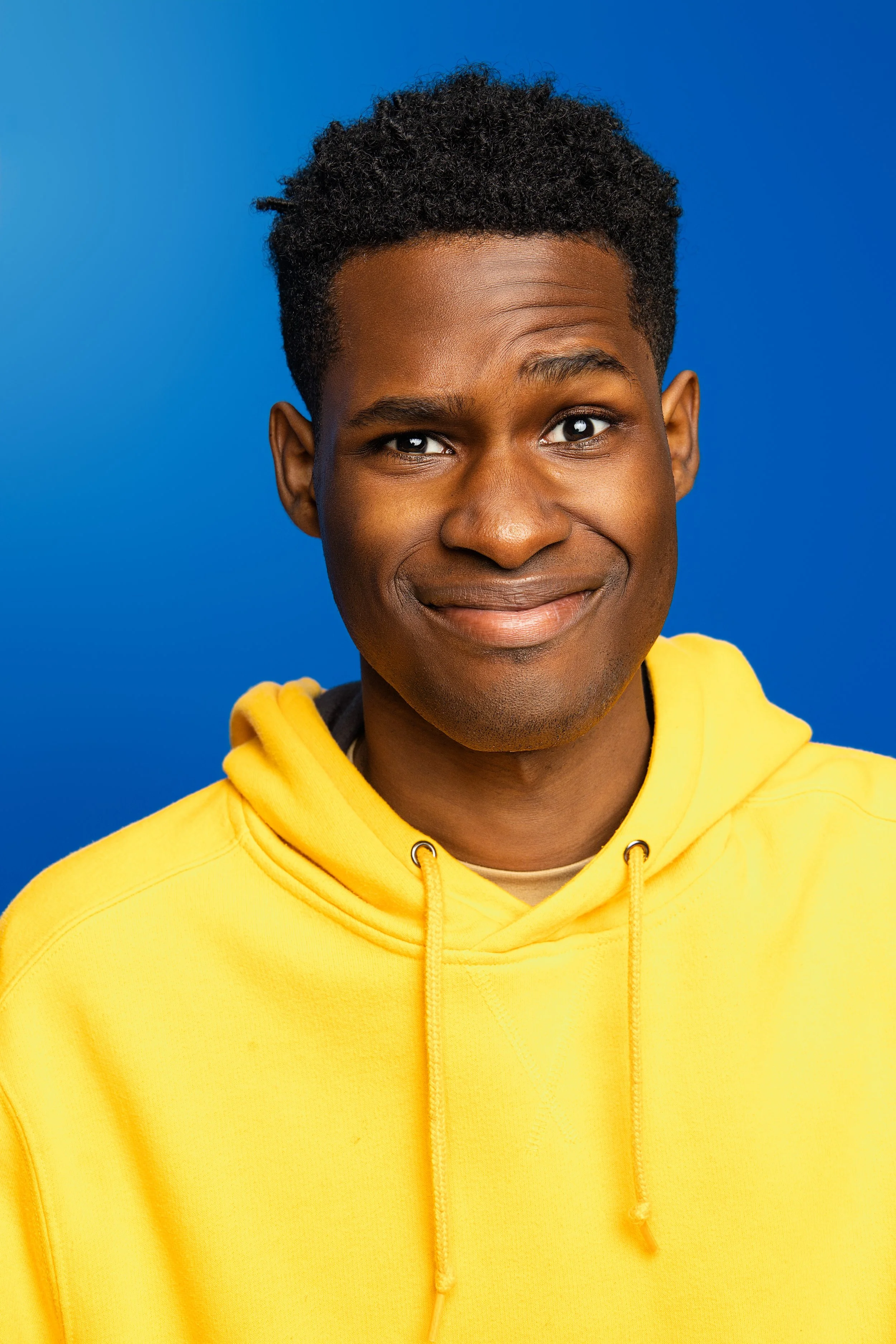 A young man with dark skin and short curly hair, smiling, wearing a yellow hoodie, against a blue background.