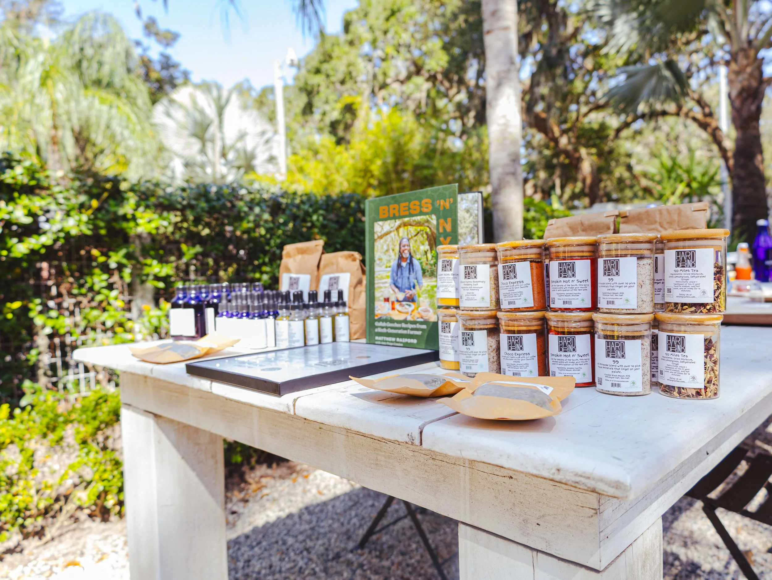 Outdoor table displaying various jars of tea and other products, with a small sign, surrounded by trees and greenery.