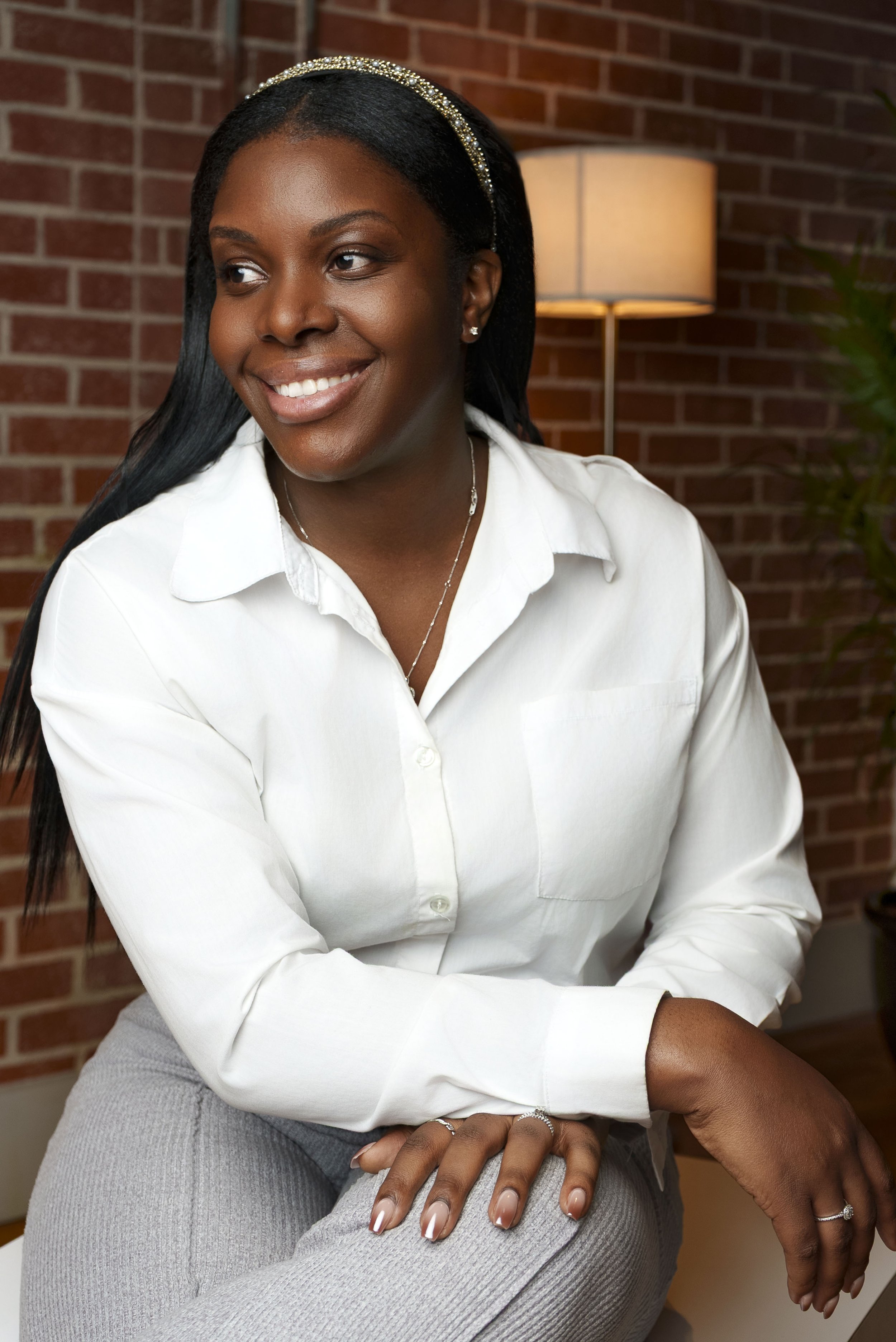 A woman with dark skin and long black hair sitting in a room with a brick wall background, smiling and looking to her left, wearing a white blouse and gray pants, with jewelry including rings, earrings, a necklace, and a headband.