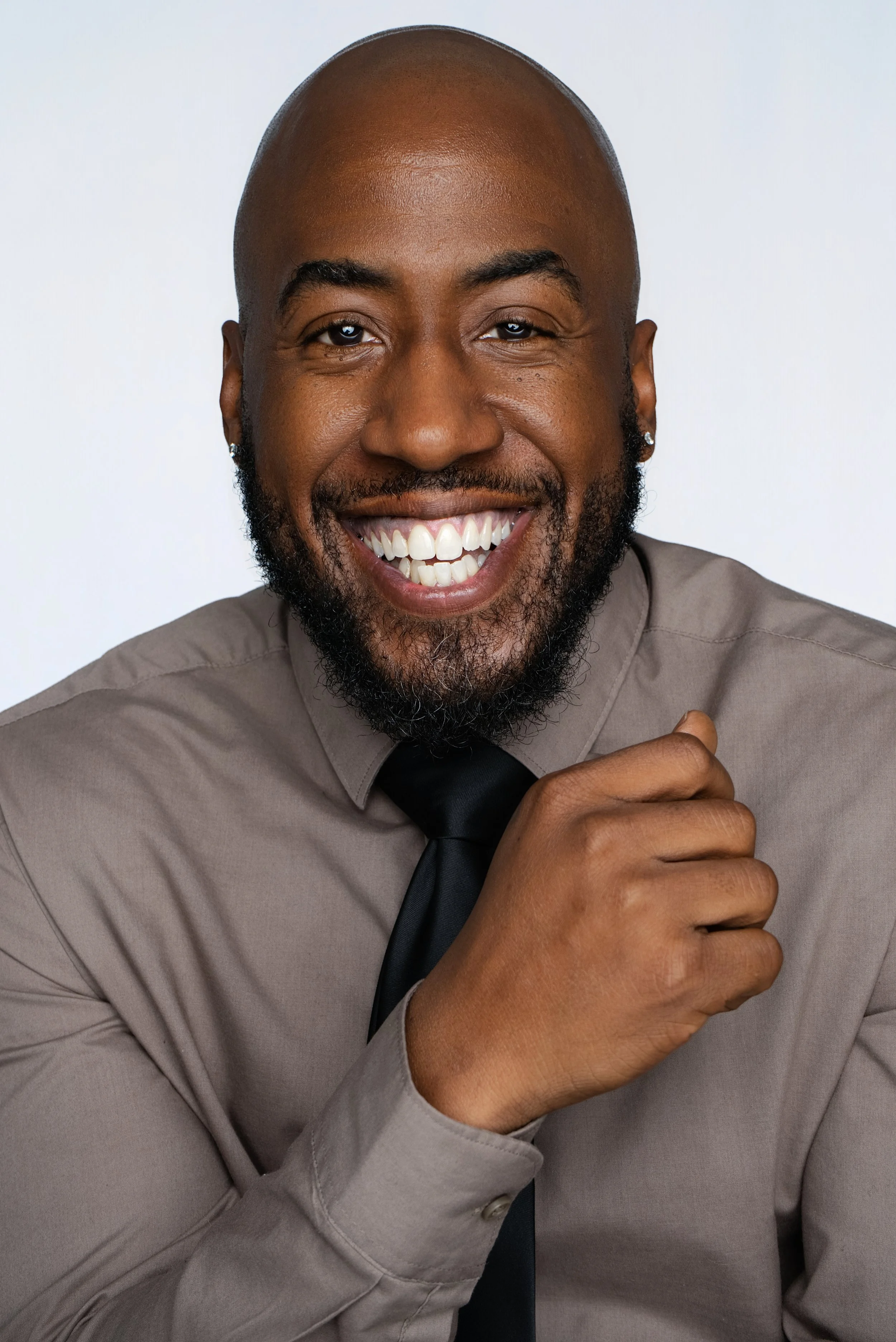 A smiling man with a beard, bald head, earrings, wearing a light brown shirt and a black tie.