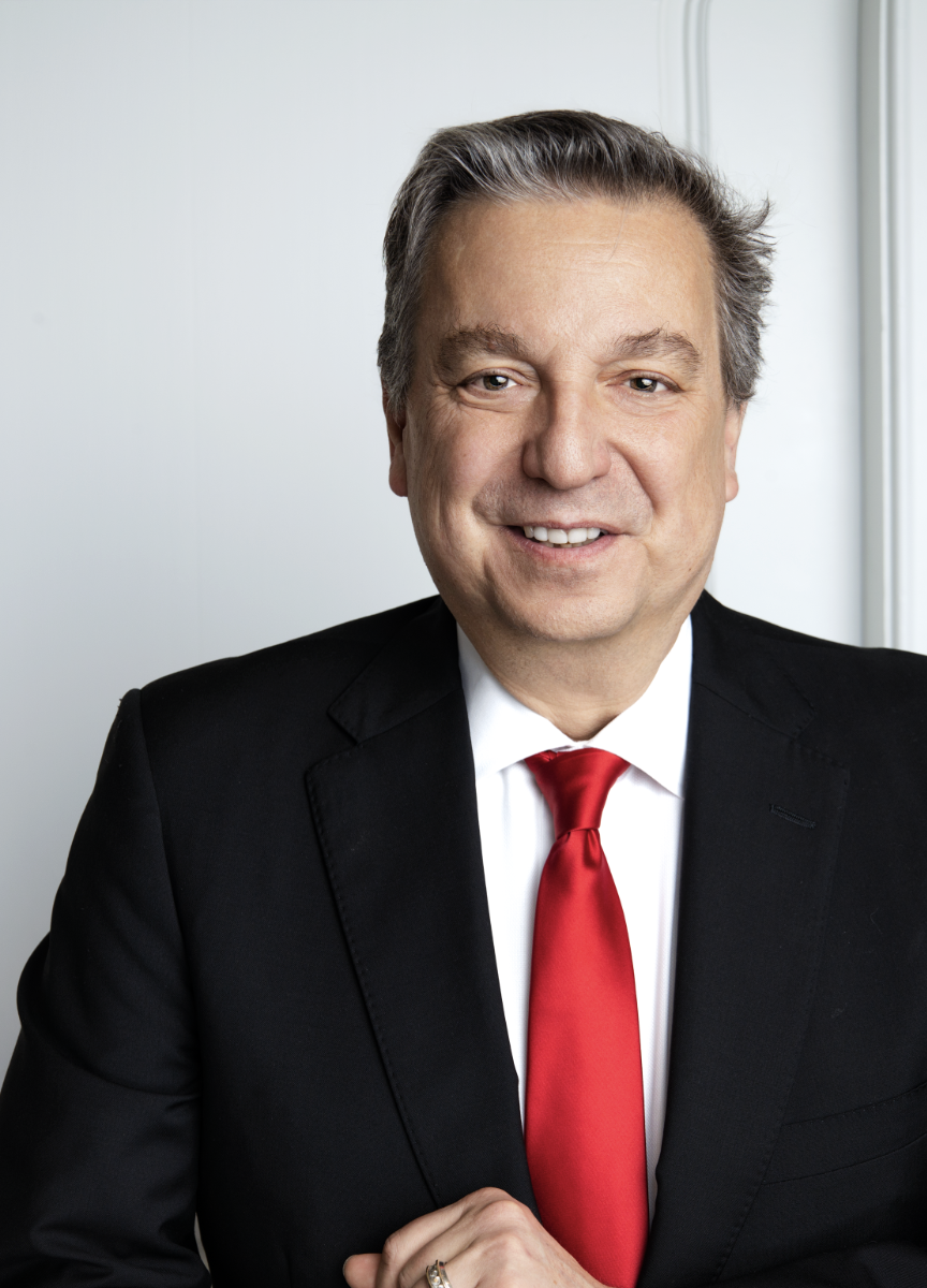 A middle-aged man in a suit and red tie smiling at the camera, standing in front of a light-colored wall.