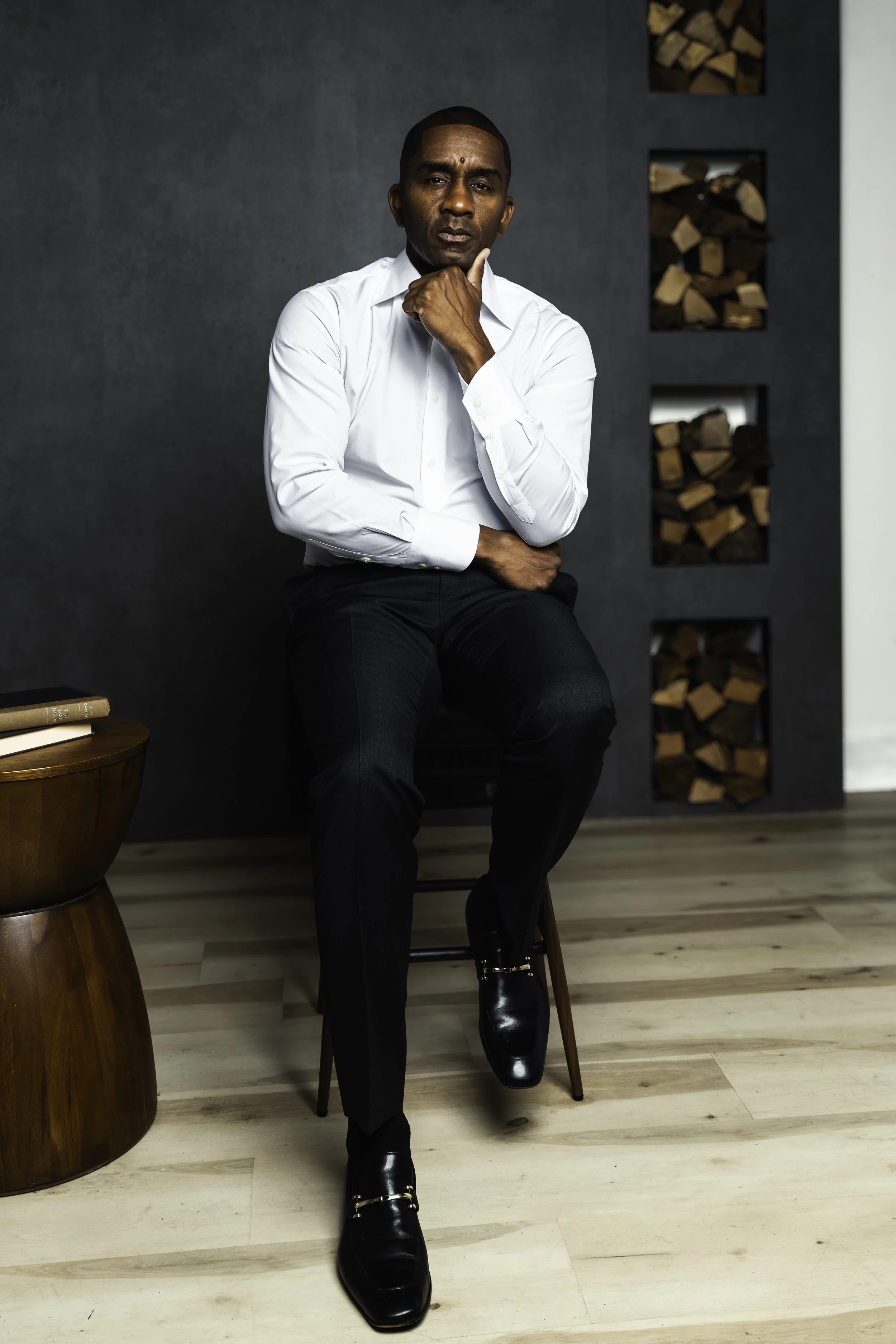 Man sitting on a chair with one hand on his chin, wearing a white shirt, black pants, and black shoes, in a modern room with a dark wall and wood logs in the background.
