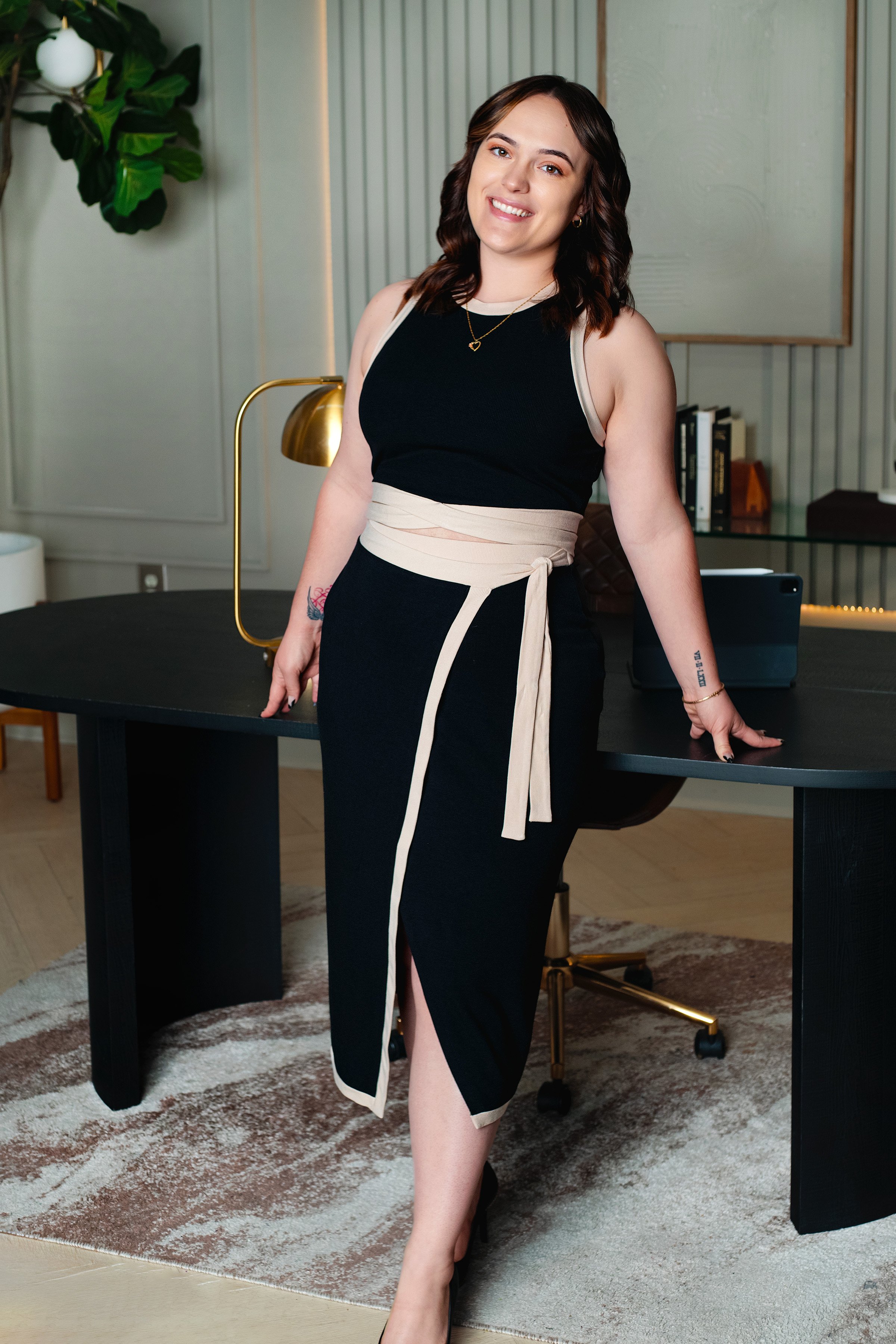 A woman with wavy brown hair wearing a black sleeveless top and black skirt with beige trim, standing in an office with a desk, gold desk lamp, and bookshelf in the background.