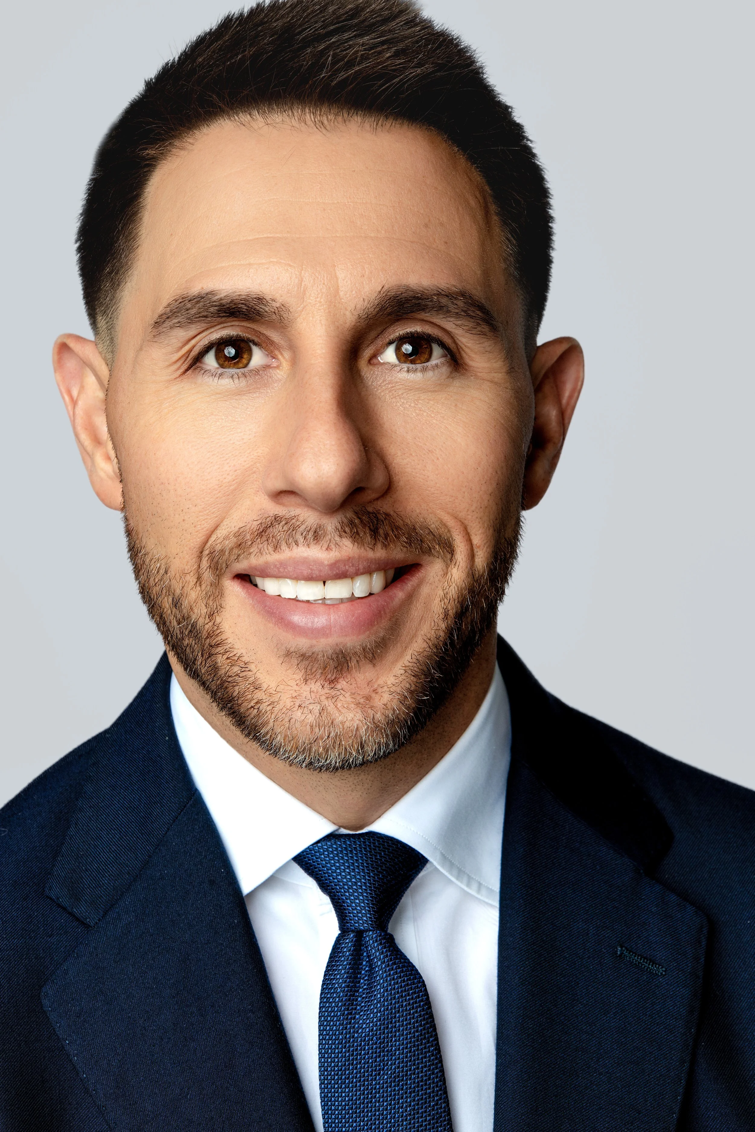 Headshot of a smiling man with brown eyes, dark brown hair, and a beard, wearing a navy suit, white shirt, and blue tie.