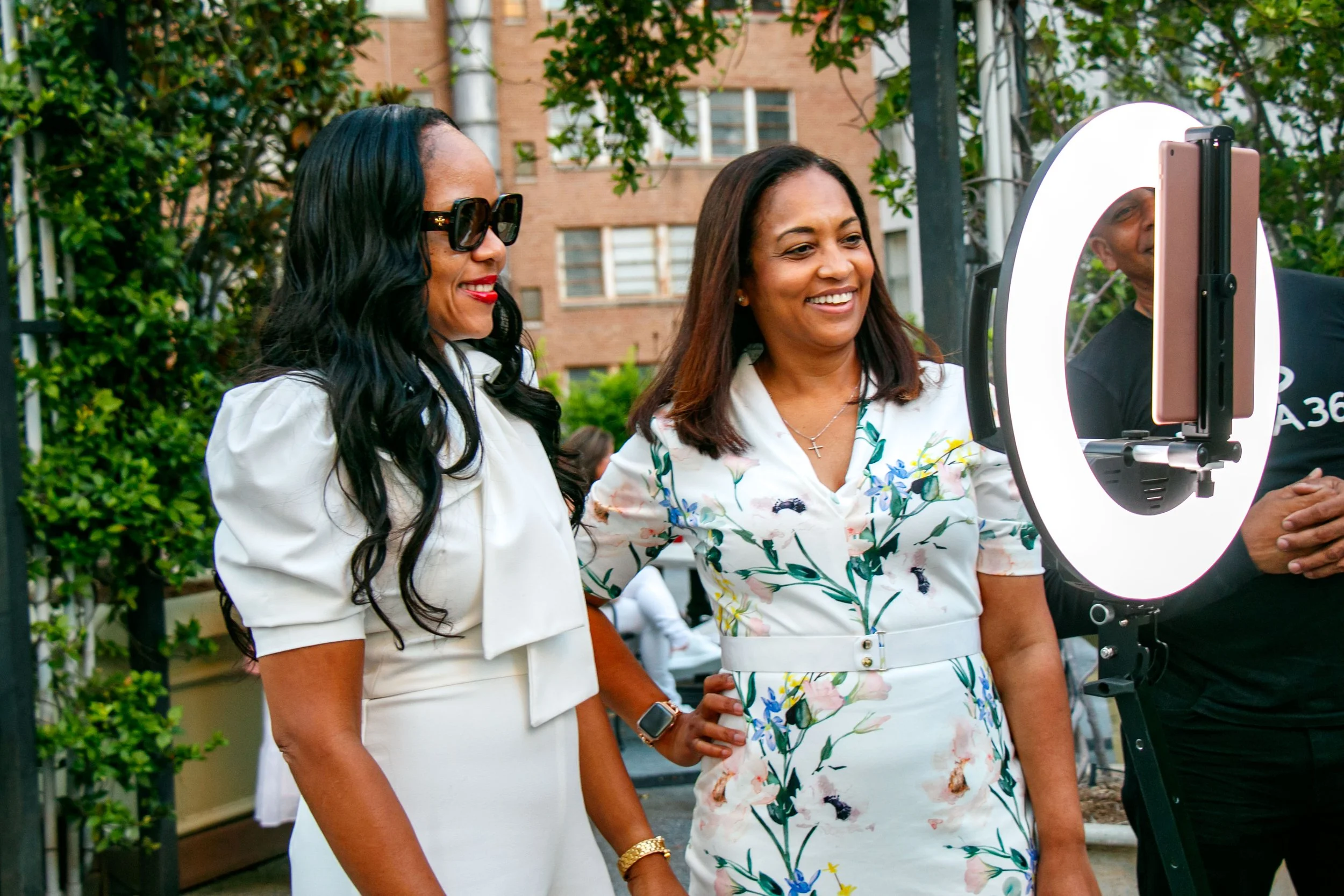 Two women standing side by side outdoors, smiling at a smartphone mounted on a ring light stand, with trees and a brick building in the background.