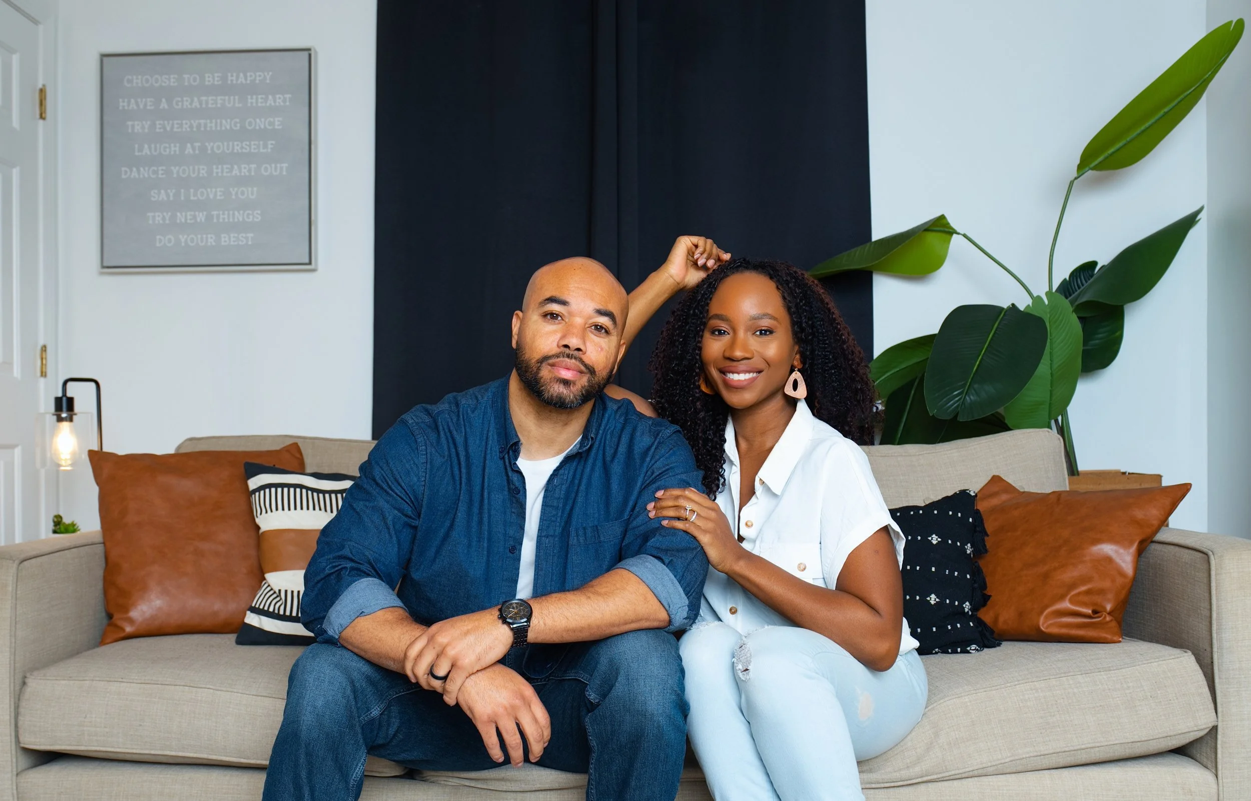 A smiling couple sitting on a beige couch with colorful pillows in a modern living room, next to a large green plant and framed wall art.