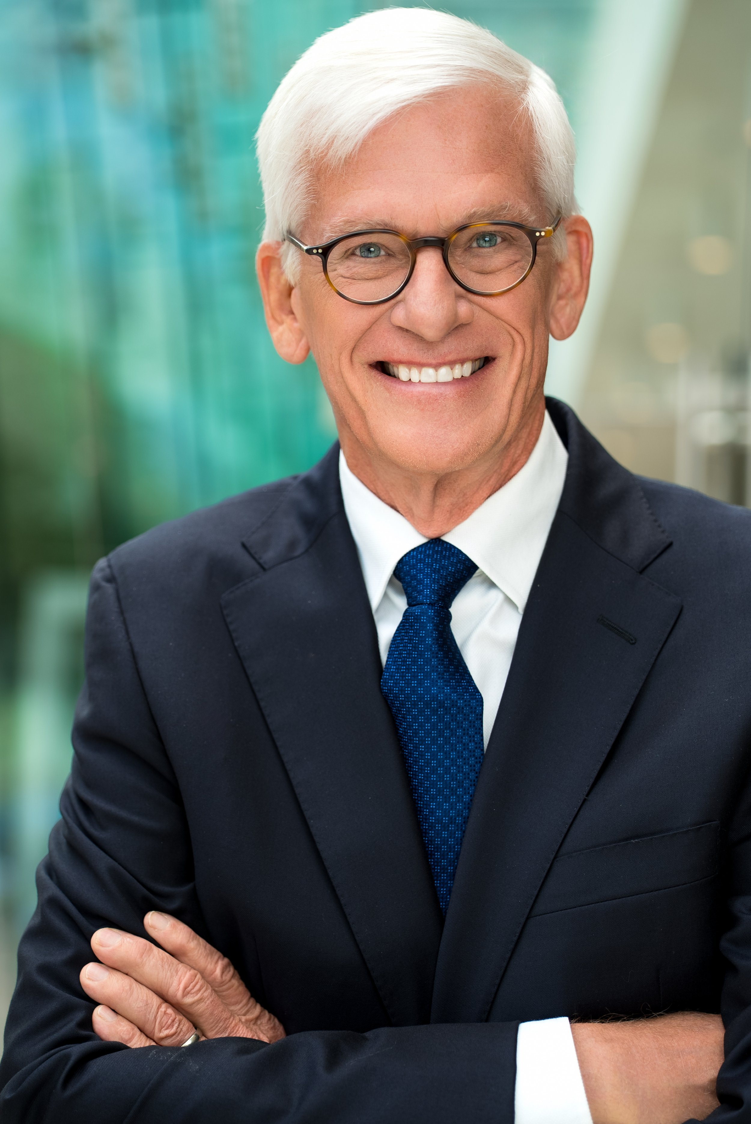 A smiling older man with white hair and glasses, dressed in a dark suit and blue tie, standing with arms crossed in front of a modern office building.