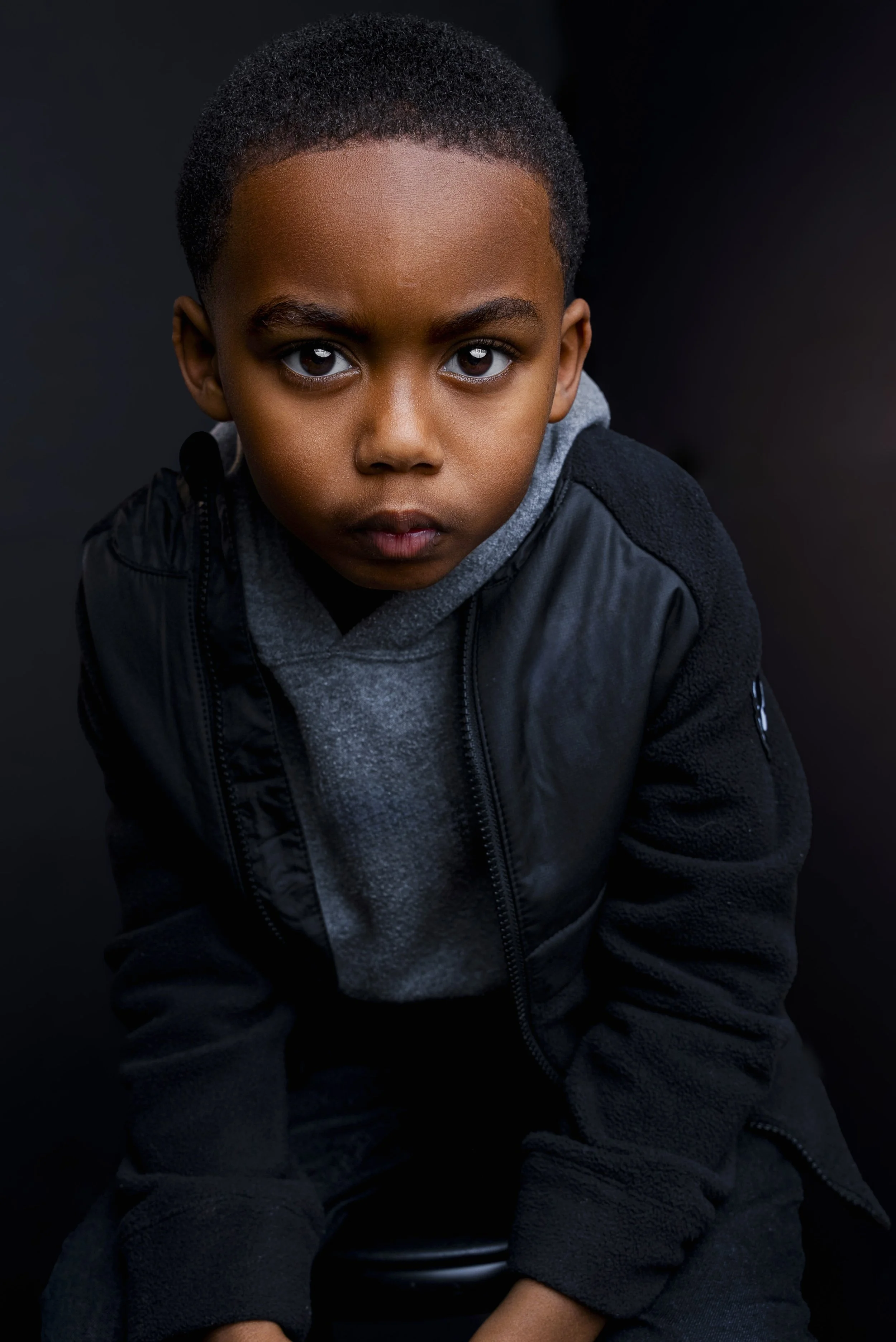 Close-up portrait of a young boy with short black hair and dark eyes, wearing a black jacket and grey hoodie, looking intensely at the camera against a dark background.