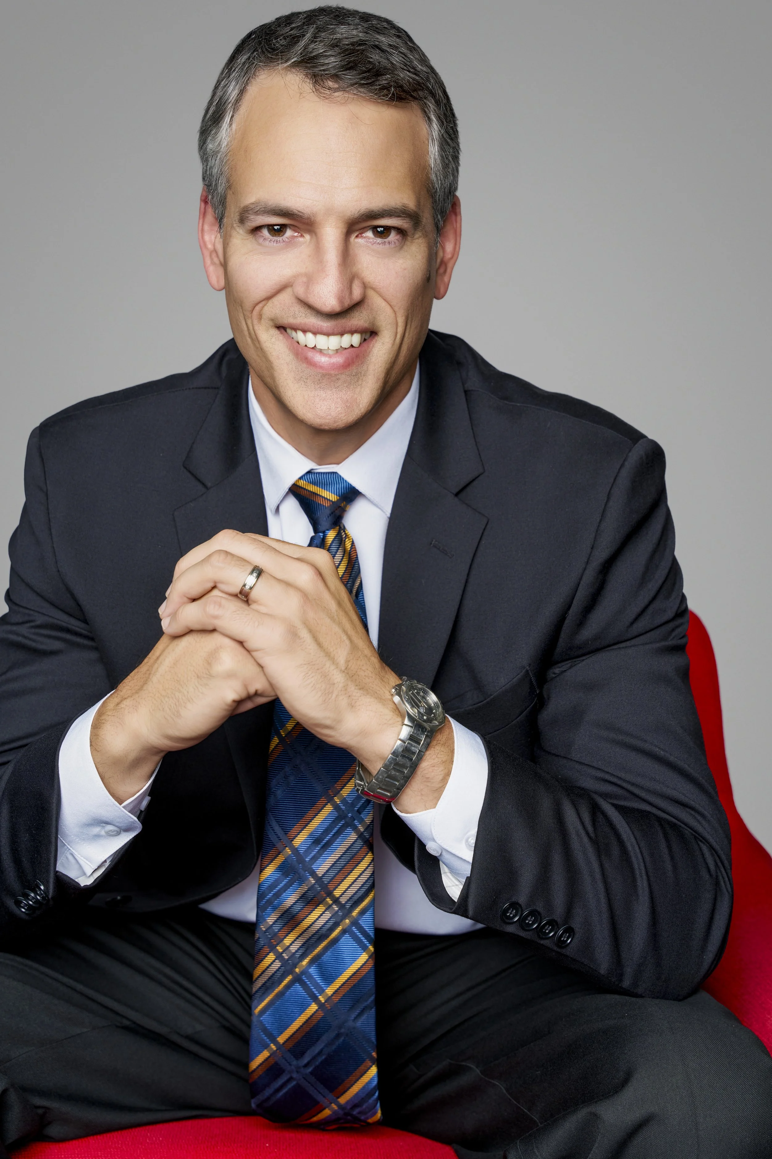 A smiling middle-aged man wearing a dark suit, white shirt, and a colorful checked tie sitting on a red chair against a neutral background.