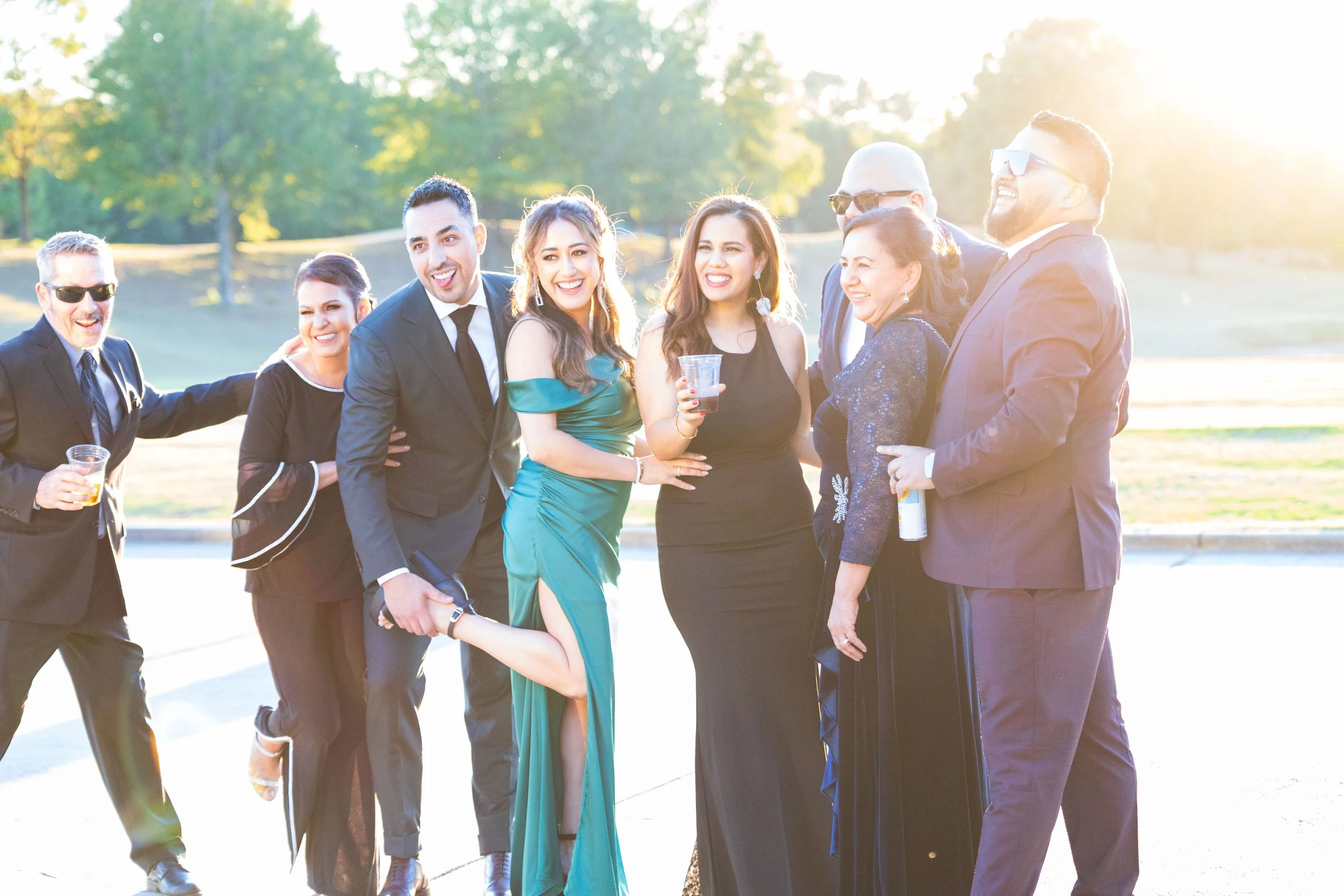 Group of people dressed in formal attire, smiling and enjoying outdoors during sunset, with trees in the background.
