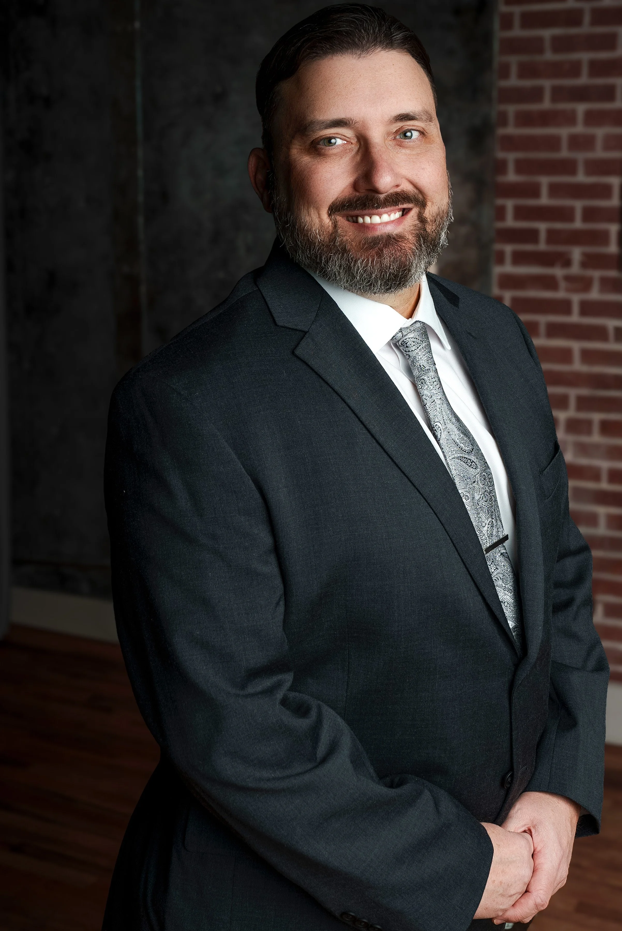 A smiling man with a beard in a dark suit, white shirt, and patterned tie, standing with hands clasped in front of him against a background of brick and textured walls.