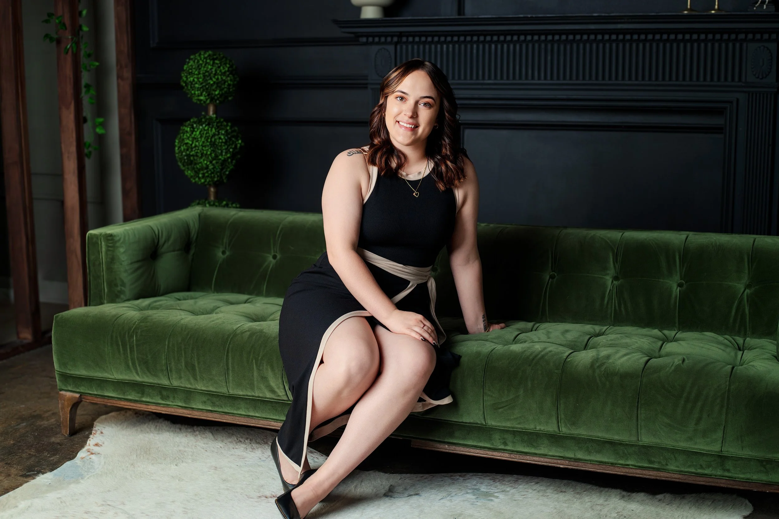 A woman in a black dress sitting on a green velvet couch in a room with dark walls and green plants.